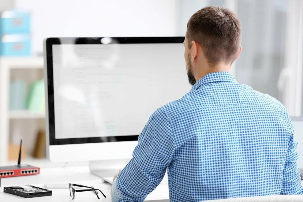 A man at a desk seen from behind, working at a monitor showing lines of code