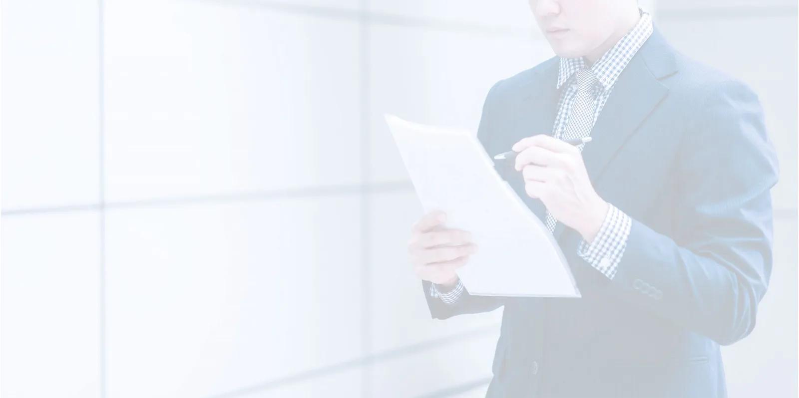 A man in a dark suit stands reading a sheet of paper in front of a bright window wall