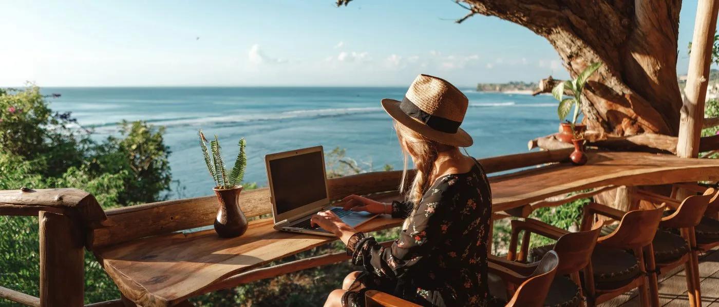 A woman in a straw hat works at a laptop on a terrace bar-top overlooking a palm-fringed coastline