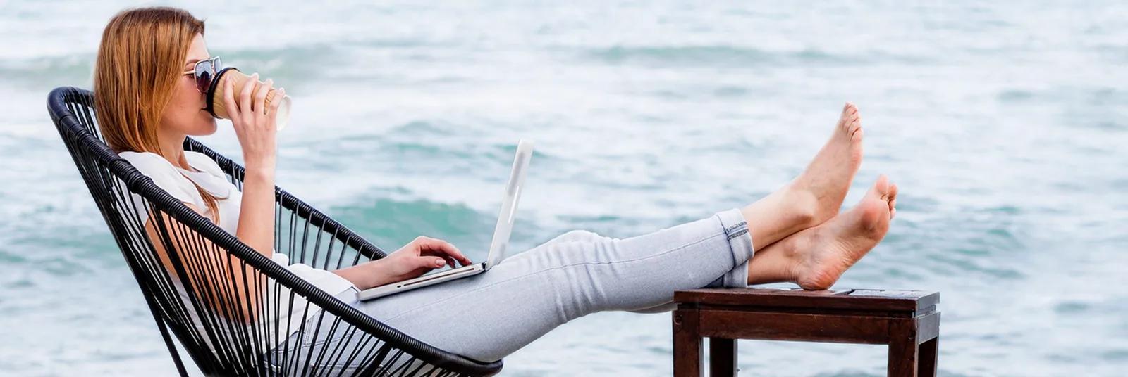 A woman reclines barefoot in a rope chair by the water with a laptop on her lap and a takeaway cup in hand