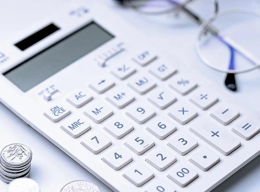 A desk calculator, a pair of glasses and a small stack of coins on a white surface