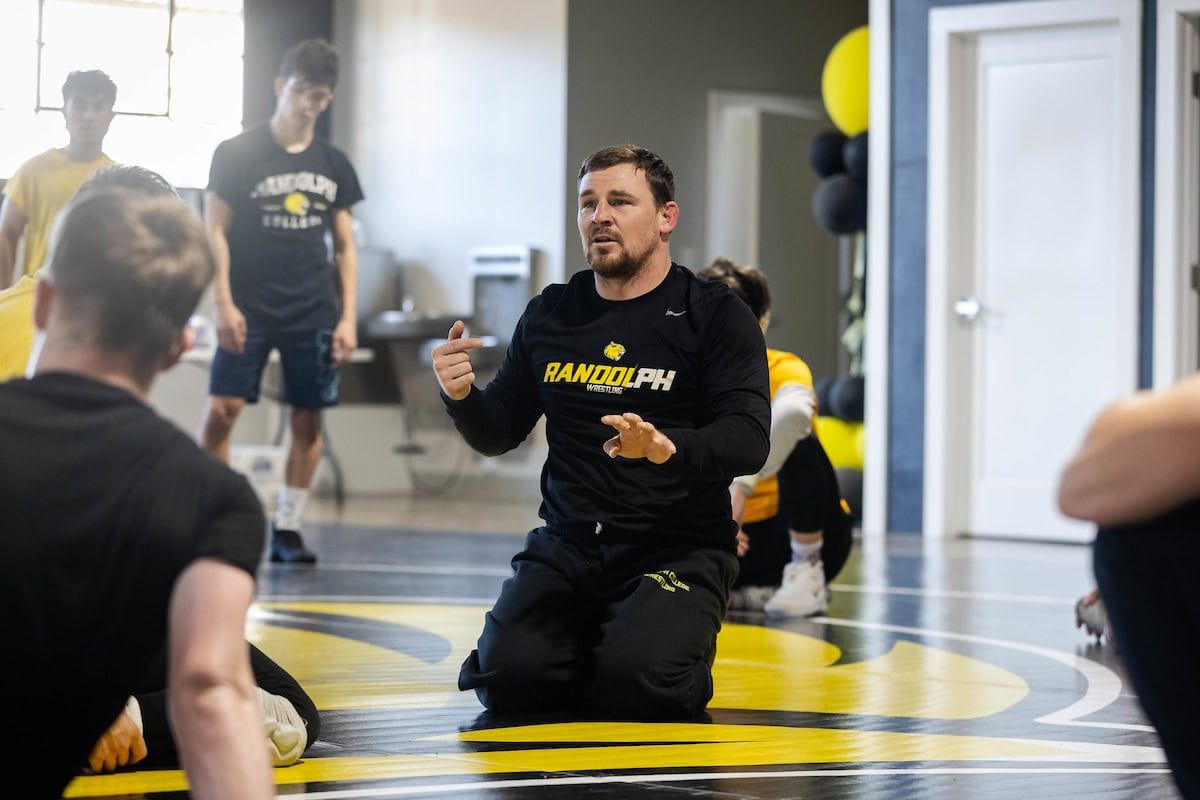 A wrestling coach leading a practice session from the mat, walking his team through the day's drilling plan.