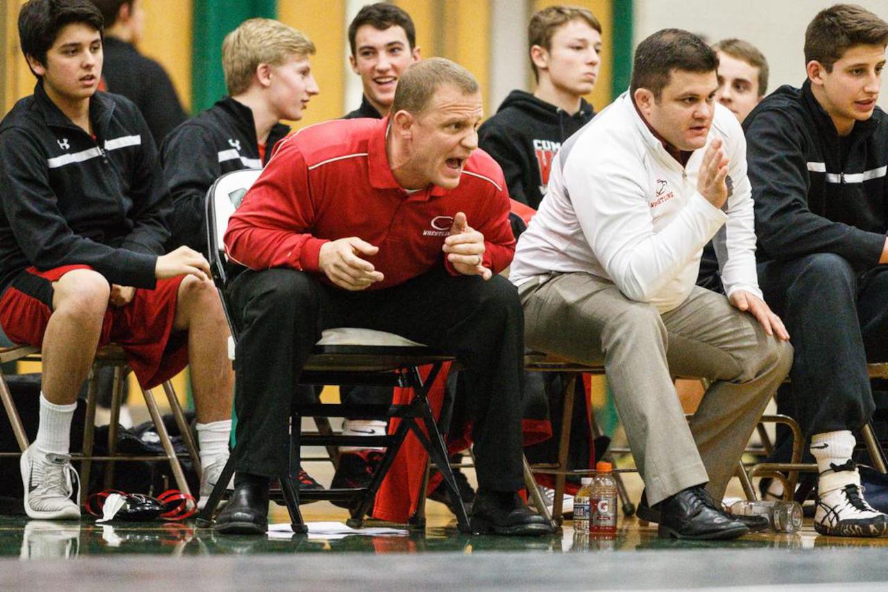 Head wrestling coach shouting instructions from the corner during a dual meet, assistant coach and team bench behind him.
