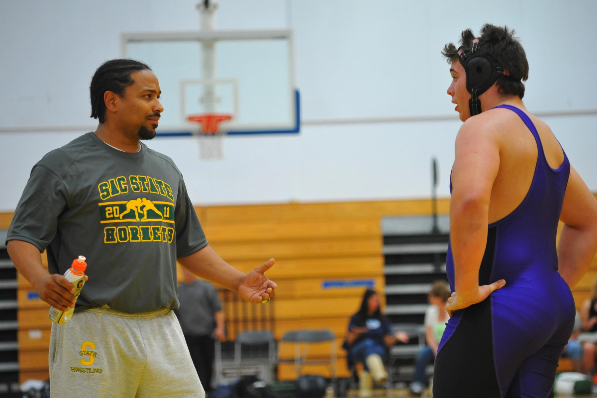 Wrestling coach in a Sac State Hornets shirt talking with a high school wrestler in a purple singlet and headgear on the side of a gym mat between matches.