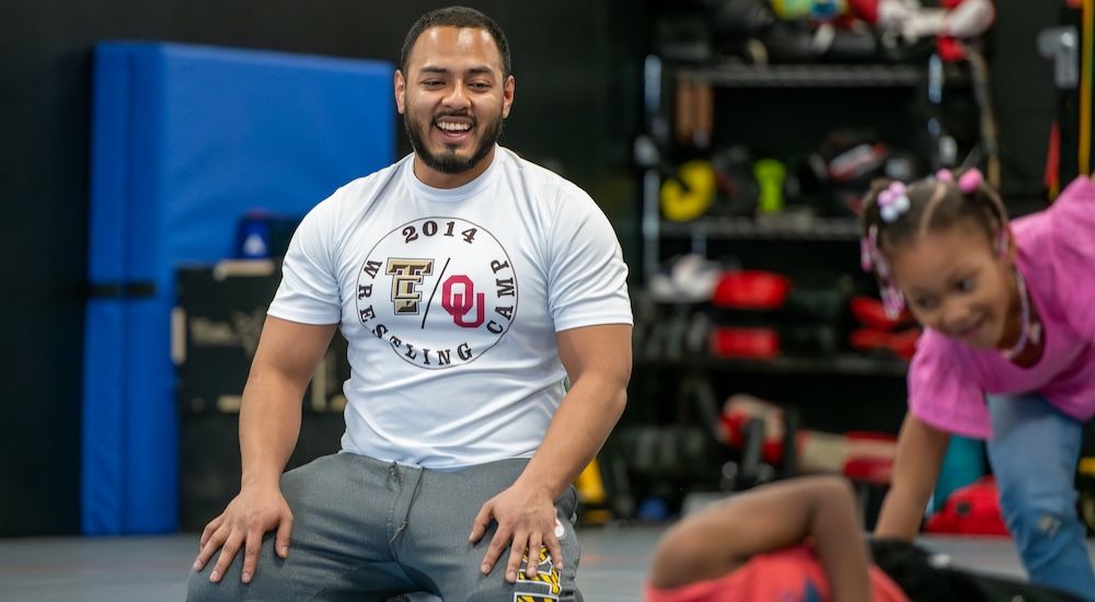 A smiling wrestling coach in an OU Wrestling Club shirt kneeling in a practice room while a young child in pink plays on the mats beside him.