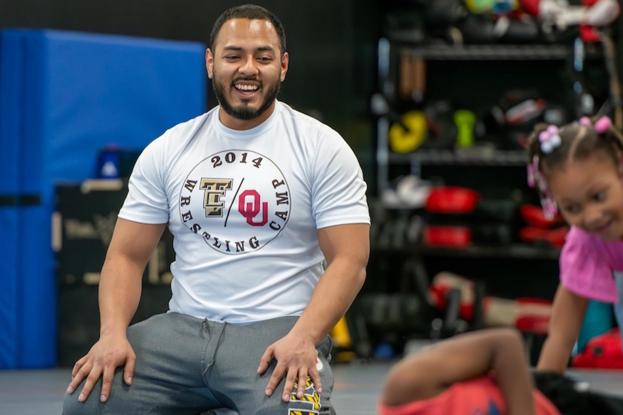 A smiling wrestling coach in an OU Wrestling Club shirt kneeling in a practice room while a young child in pink plays on the mats beside him.