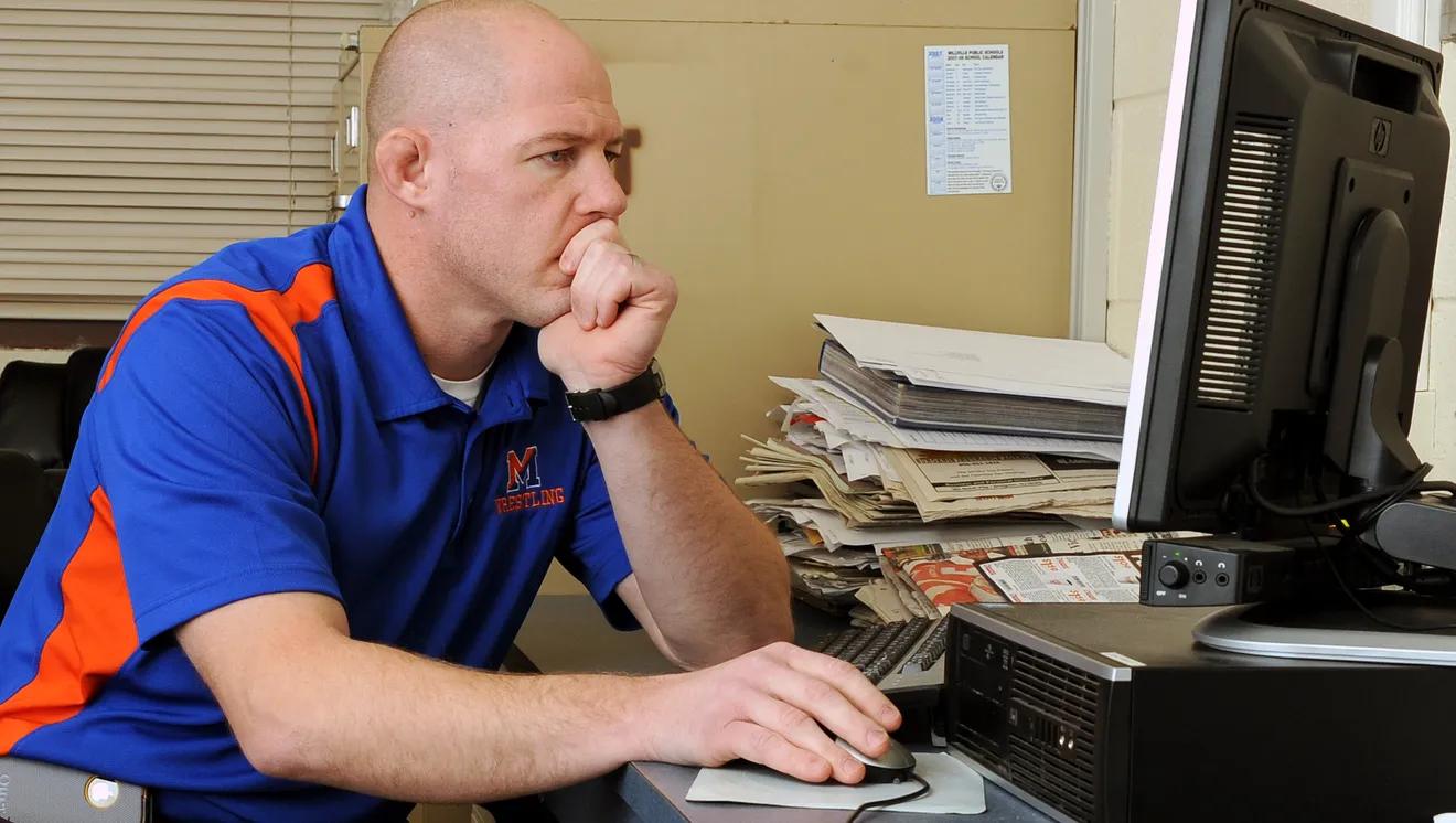 Wrestling head coach in a school polo working at his office desktop computer, with stacked binders and program paperwork beside the monitor.