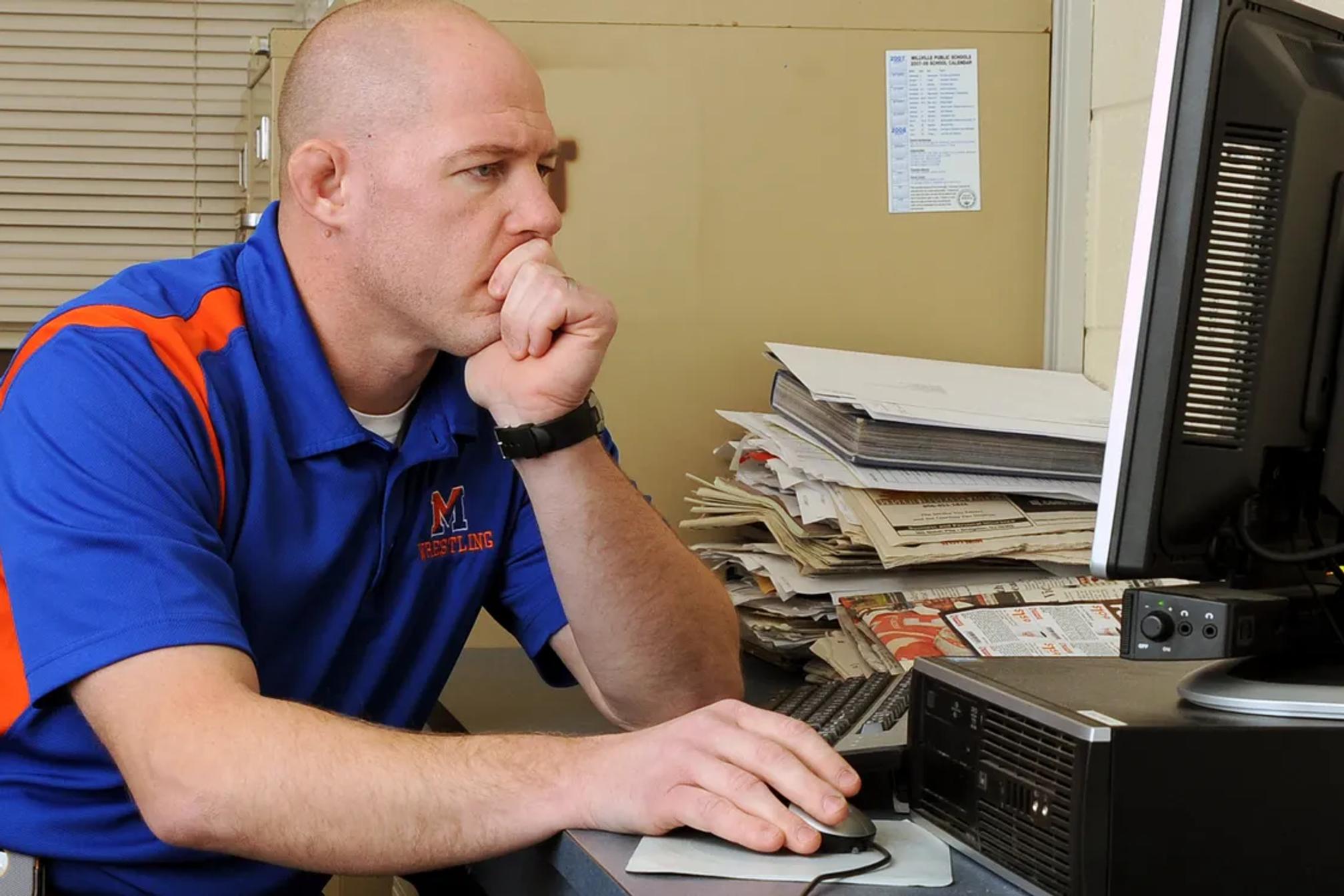 Wrestling head coach in a school polo working at his office desktop computer, with stacked binders and program paperwork beside the monitor.
