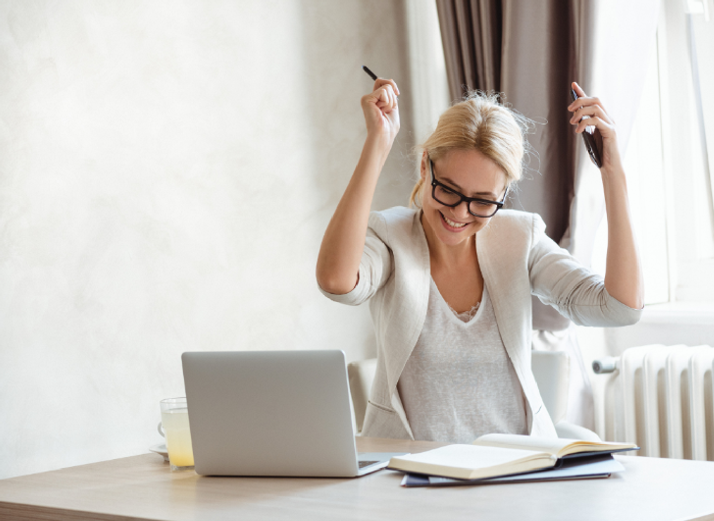 blonde woman wearing glasses and working from home with laptop, books and phone, celebrating with hands raised in the air