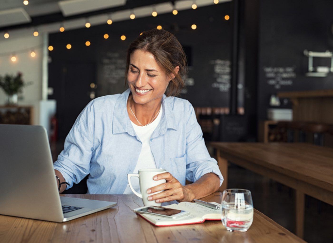 woman happily working on a laptop in a cafe with coffee and fairy lights