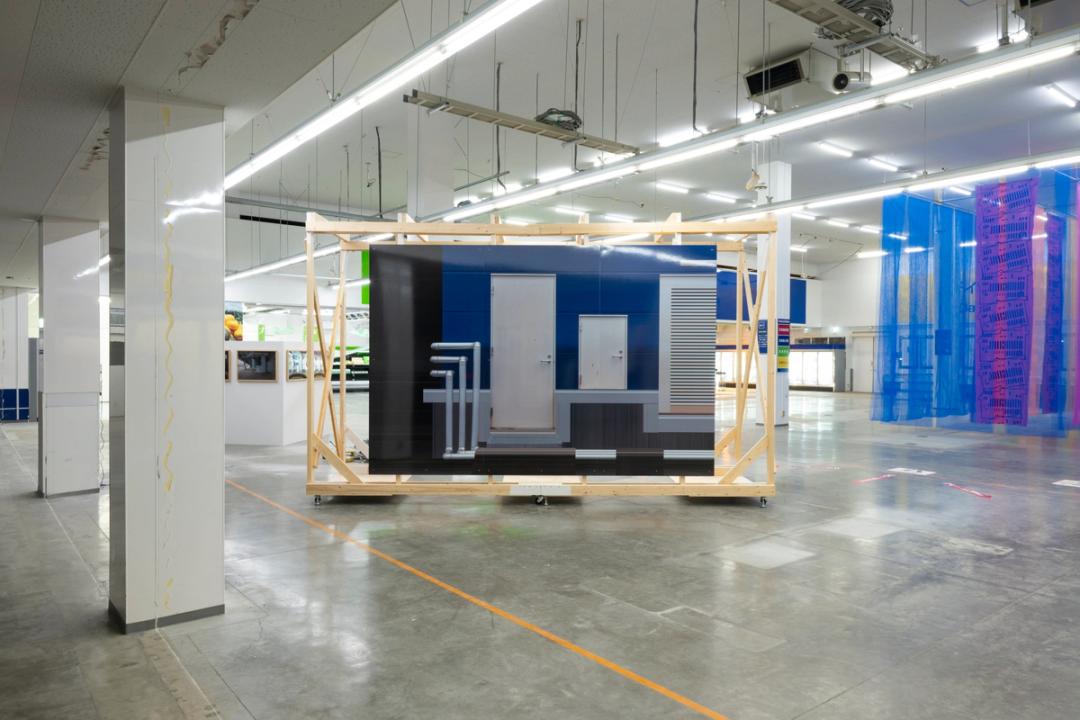 《再構築された壁》に展示された《壁と扉》，2025　かつて商業施設だったBLUE CUBEは、教育施設へと転用され、採光確保のため間仕切り壁が撤去された。藤原禎之はこの改修工事を取材し《 3枚の記録写真》に収め、間仕切り壁の廃材を再利用して2種の仮設壁《再構築された壁》と《WHITE CUBE》を制作した。元の部材をほぼそのまま組み直して作品化する方法は、既存建築の構造を活かして用途変更を 行う「コンバージョン建築」と共通する。 　構造が剥き出しの《再構築された壁》は車輪が付いた可動壁だ。特定の位置にとどまらない壁は、常に「仮定・保留」され、建築の変化のプロセスを体現する。BLUE CUBE裏口を写した《壁と扉》は、ドアの向こう側にある空間を想像させる。写真にはピクセルを引き伸ば す微細な編集が施され、藤原が「意味のキャンセル」と呼ぶ抽象化が見られる。扉や配管以 外のディテールがノイズとして処理され、建築のイメージに普遍性と解釈の余地が同時に生 まれてくる。（文：渡辺 眞弓 多摩美術大学美術館 学芸員）　photo：©️齋藤 彰英