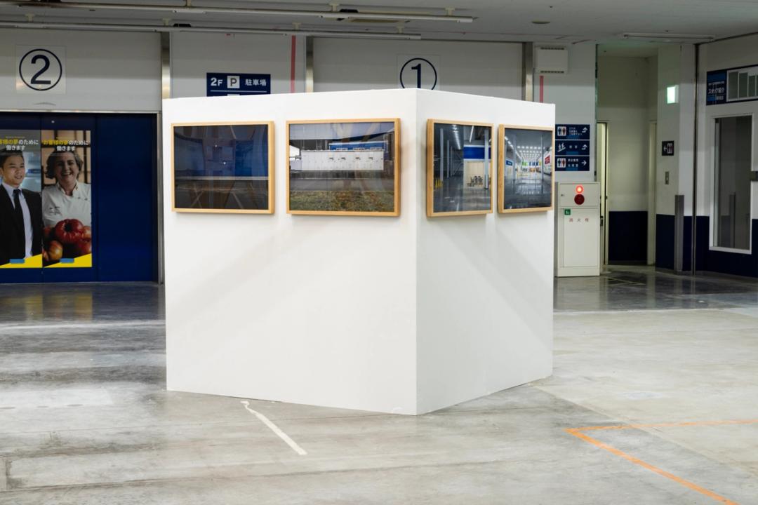  《WHITE CUBE》に展示された《 8枚の記録写真》部分，2025　《WHITE CUBE》は美術館の展示空間を外部へ反転させ「記号化」したスタティックな作品だ。撤去された壁の石膏ボードが使われ、ビス穴の補修跡が時間的厚みを付与している。そこに均等に配置された《 8枚の記録写真》はニュートラルな展示形式を踏襲し、建築の人工的な造形美を鑑賞対象として提示する。当初は既存の間柱の高さ3mの壁を想定し たが、設営中に182cmの「身体スケール」へ縮小された。この小さな箱は、巨大なスーパー マーケット跡地の中で異質な存在感を放ち、作品に価値を与えてきた美術館の権威や制度性を相対化する。これは展示空間のあり方を再考させる「ポスト・ホワイトキューブ」 ※の表明とも解釈できる。（文：渡辺 眞弓 多摩美術大学美術館 学芸員）　photo：©️齋藤 彰英