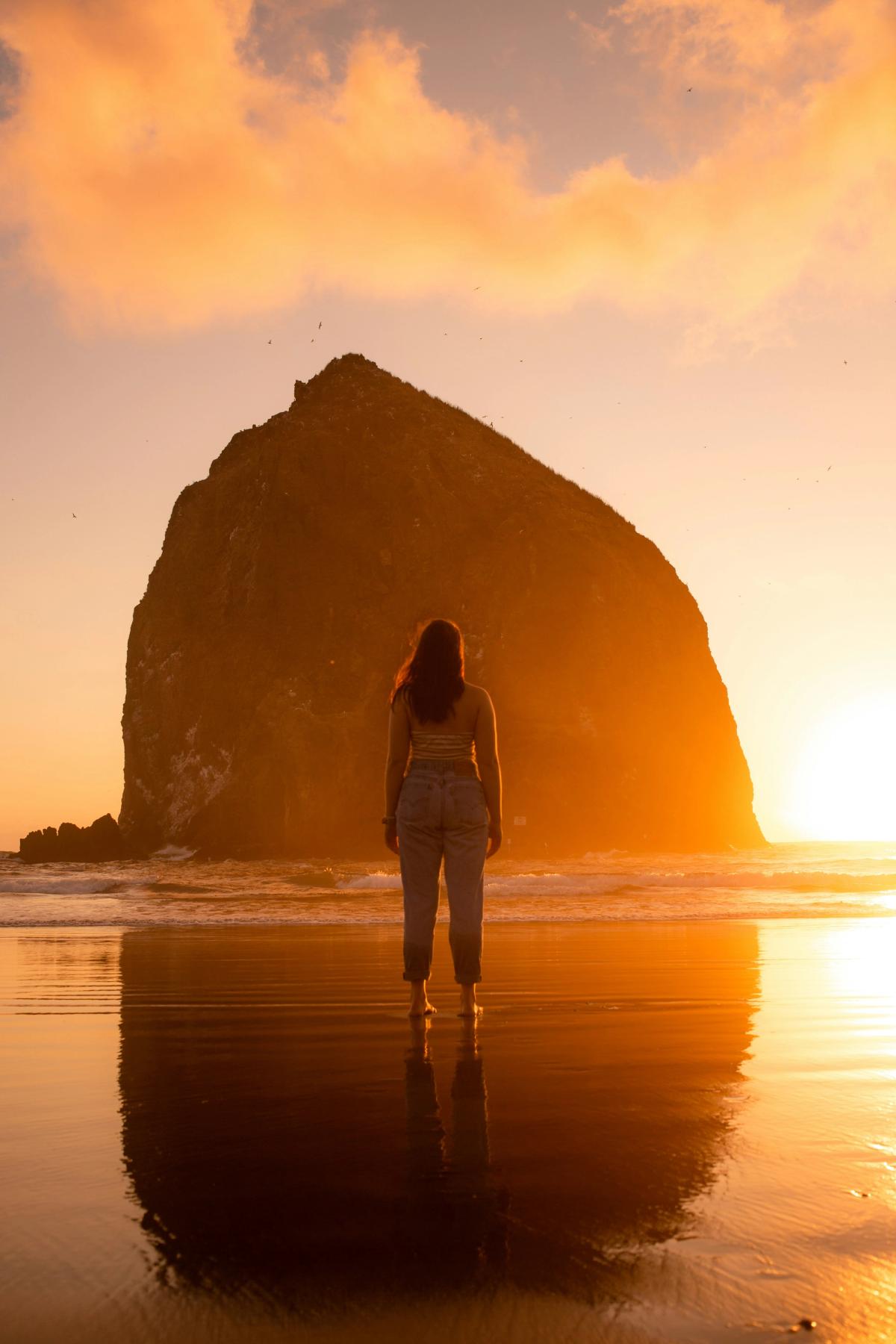 Woman stands on a beach at sunrise looking out, the sun peeking from behind a tall island