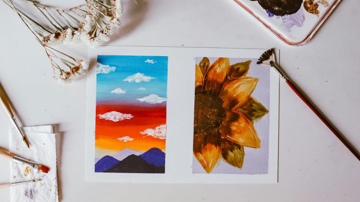 A painting in progress; brushes and dried flowers surround the paper on a plain desk.