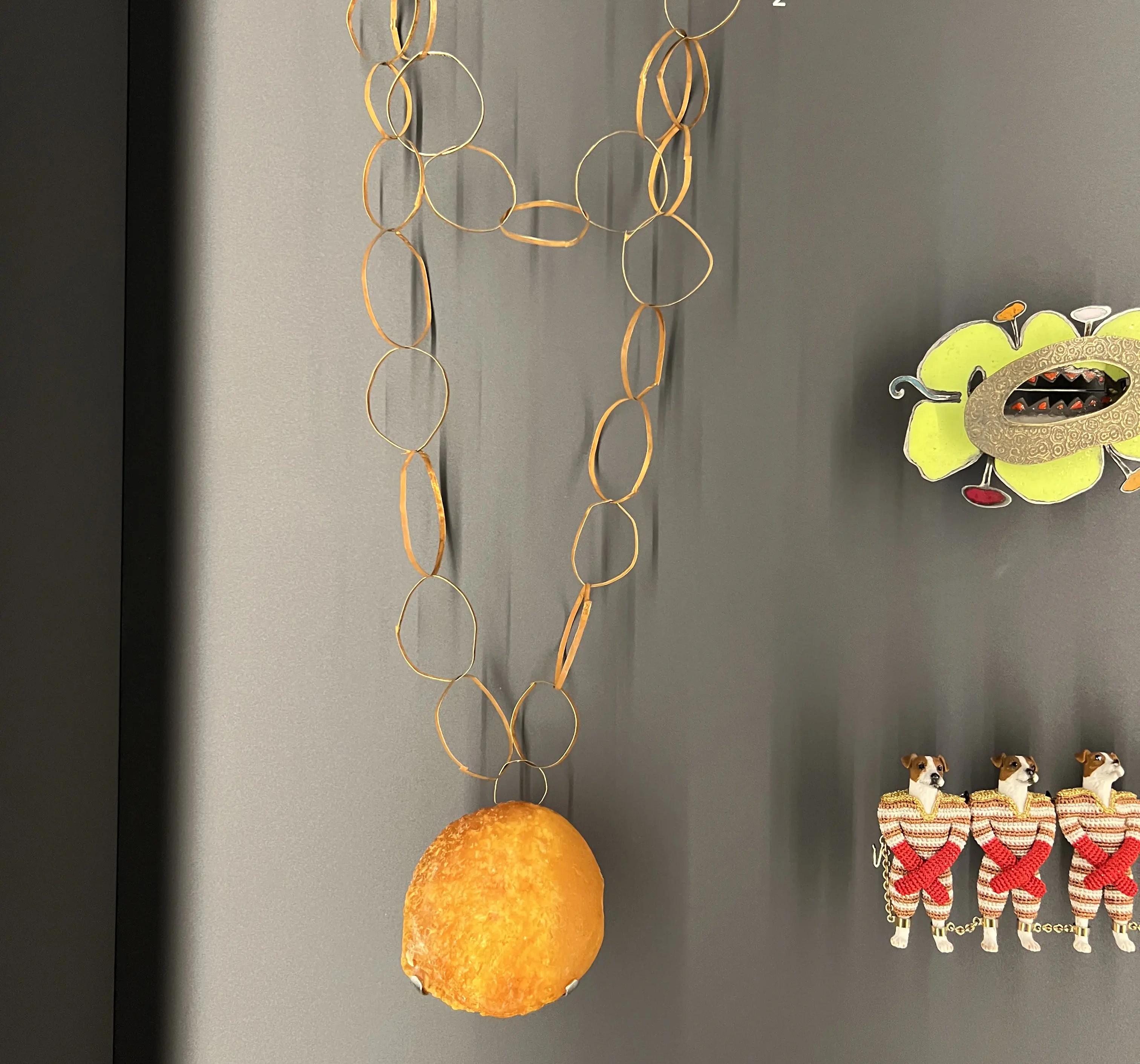 Smykkelignende installasjon av tynne ringer med en stor gul form nederst, vist mot en mørk vegg.