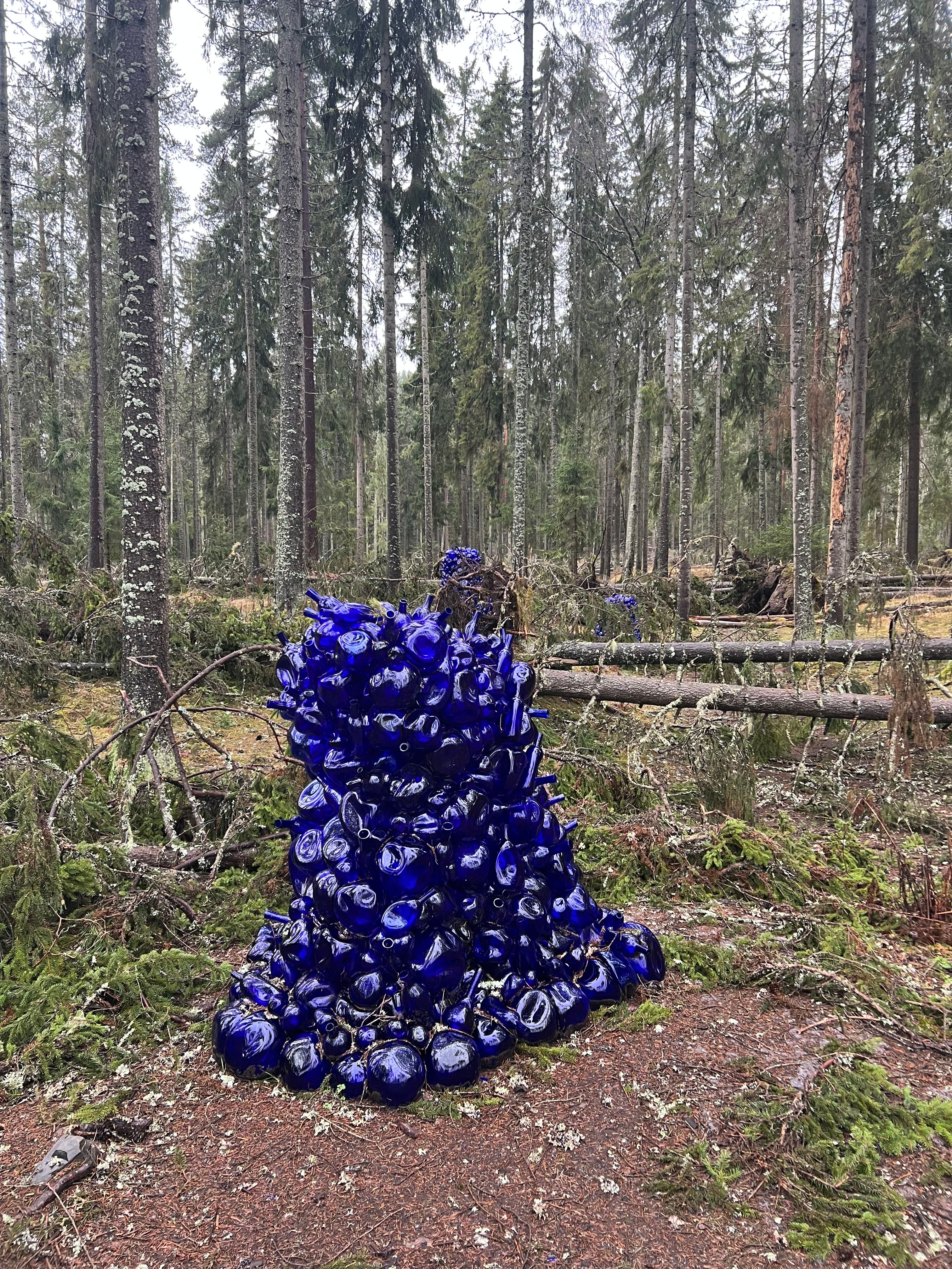 Foto av en skulptur laget av sammenpressede flasker i koboltblå glass. Dette er én av 6 skulpturer som står i skogen på Kistefos museum. Du skimter 2 skulpturer til i bakgrunn av bildet.