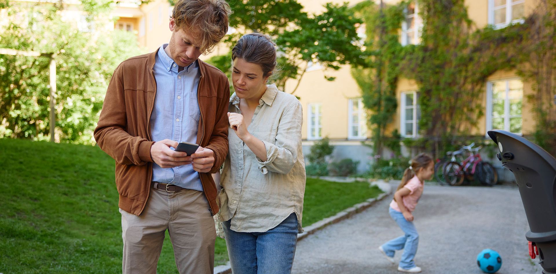 Par som ser på mobiltelefon mens barnet spiller fotball i bakgrunn.