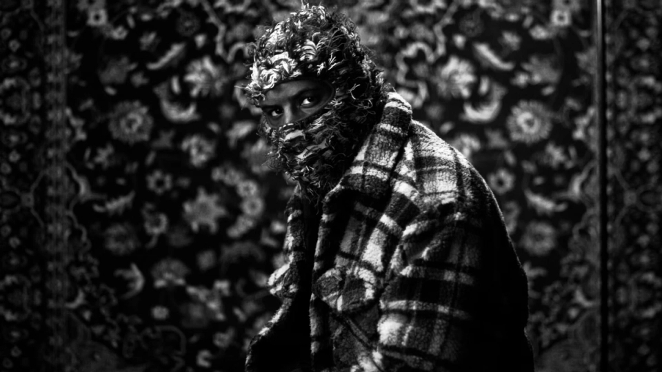 Close-up of Patrik Kabongo in a textured mask and plaid jacket, set against a patterned backdrop in the “Northface” music video.