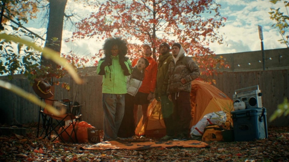 A group of friends stands together in a backyard campsite surrounded by autumn leaves, with a bright orange tent and camping gear behind them.