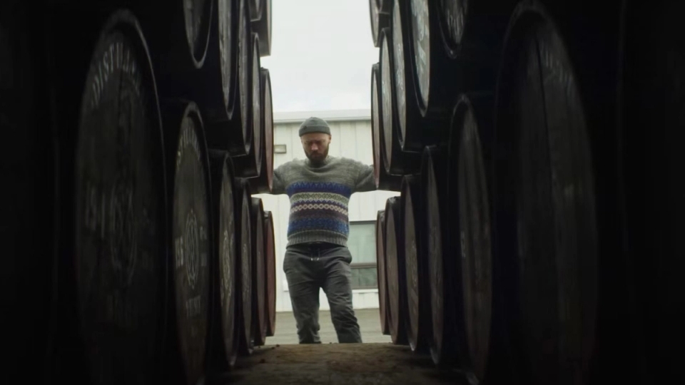 Man standing between rows of stacked whisky barrels in an open-air distillery corridor.