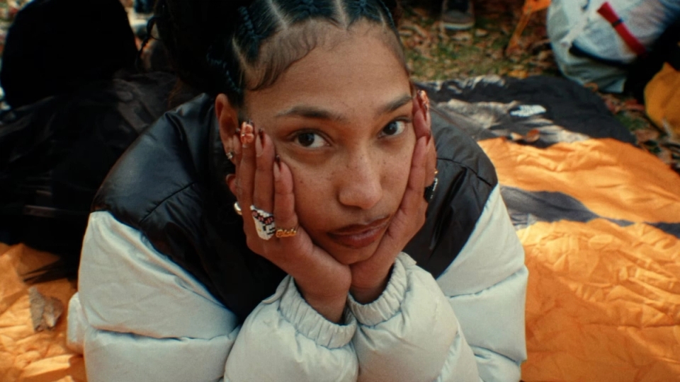 A young woman rests her face in her hands while lying on a sleeping bag outdoors, framed by fall leaves and soft light.