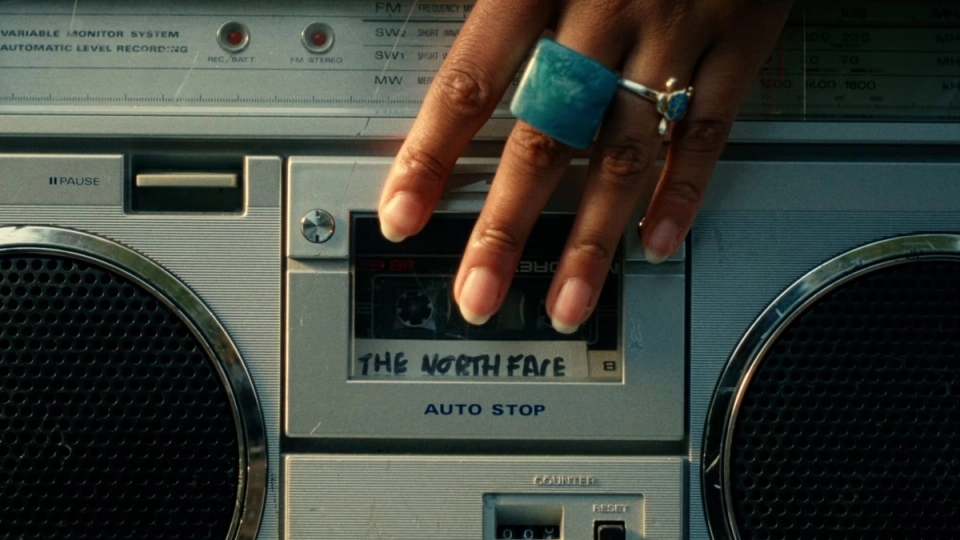 A hand with turquoise and silver rings presses play on a retro boombox cassette labeled “The North Face.”