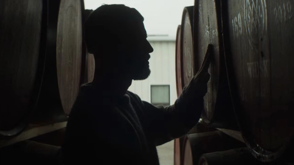 Silhouette of a person inspecting large wooden barrels inside a distillery’s aging room.