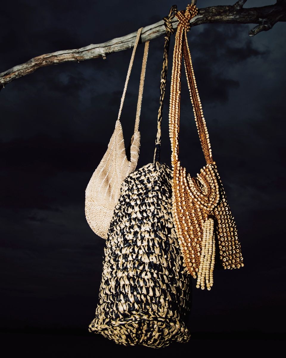 Three beaded and woven handbags hanging from a branch against a dark backdrop