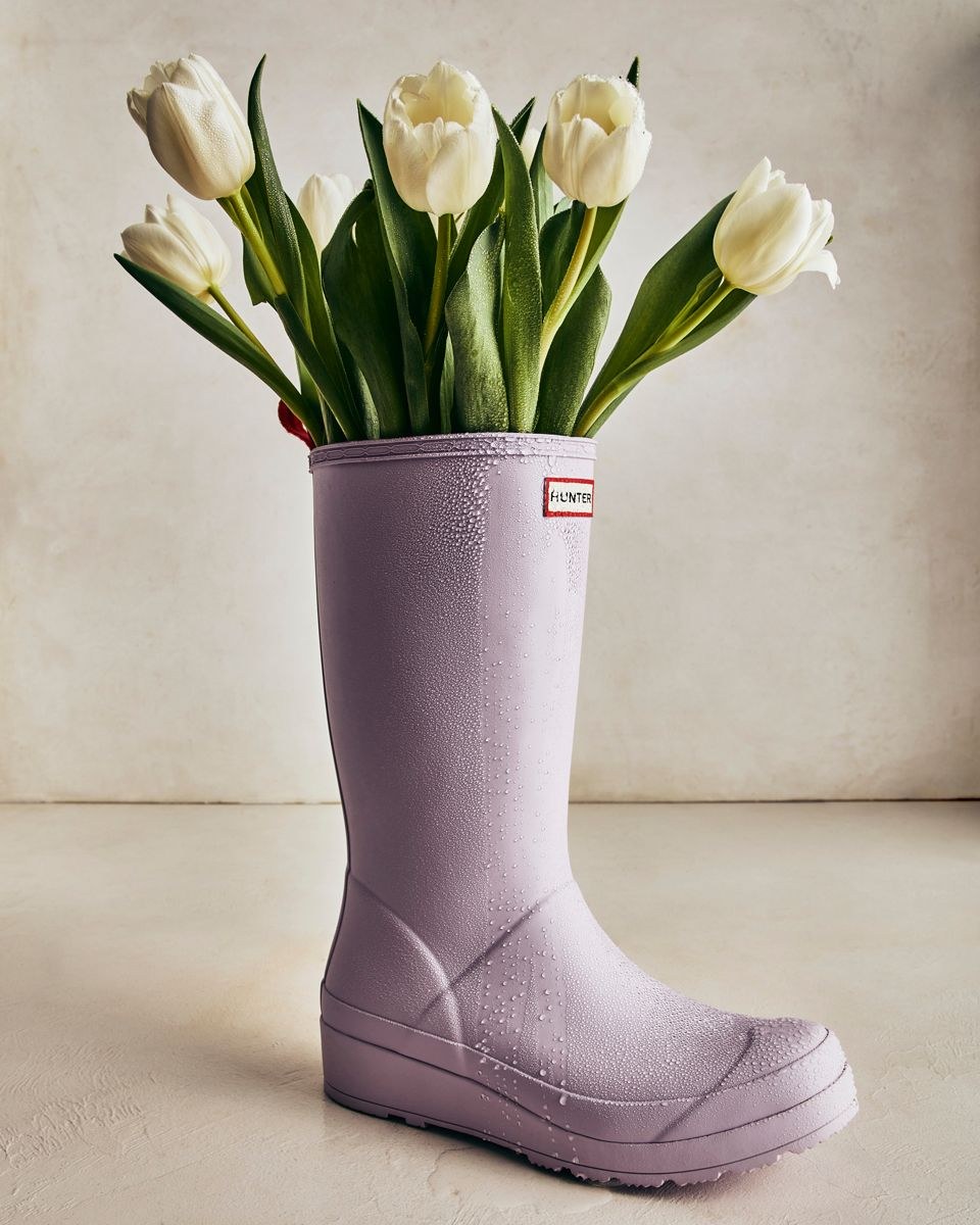 A lavender rain boot holding white tulips on a pale studio set