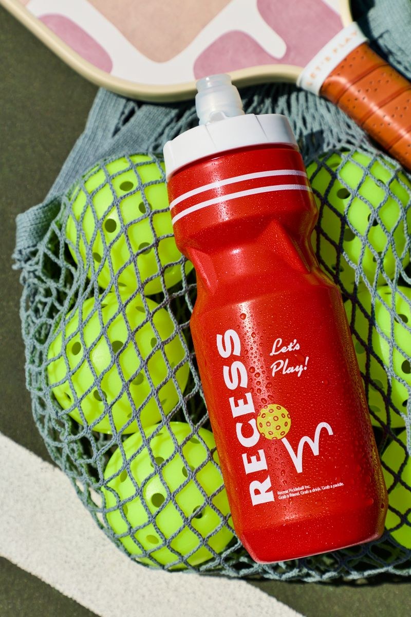 A red water bottle arranged with pickleballs, paddles, and a mesh bag