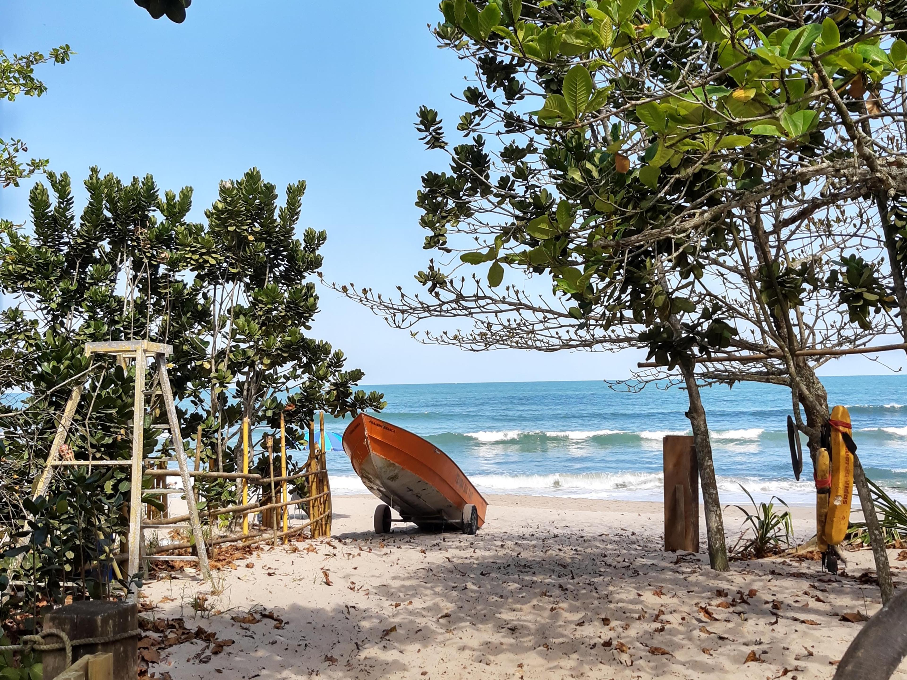 Foto tirada da areia na Prainha Branca (Guarujá). Pode-se ver também algumas árvores, um barco, e o mar ao fundo.