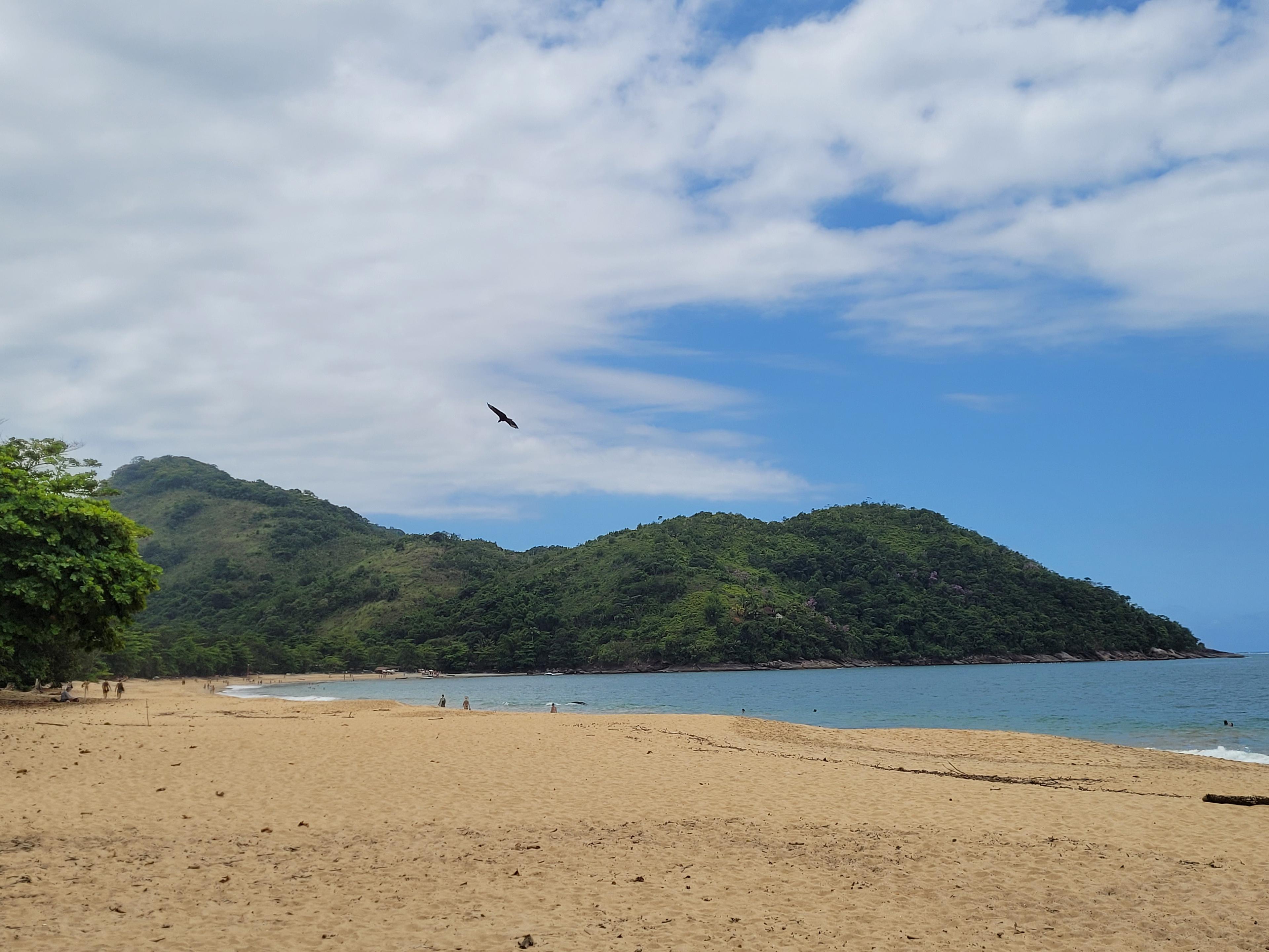 Foto tirada da areia na Praia Grande do Bonete, em Ubatuba. Pode-se ver também o mar, um morro no canto da praia, e algumas nuvens no céu contrastando com o céu azul.