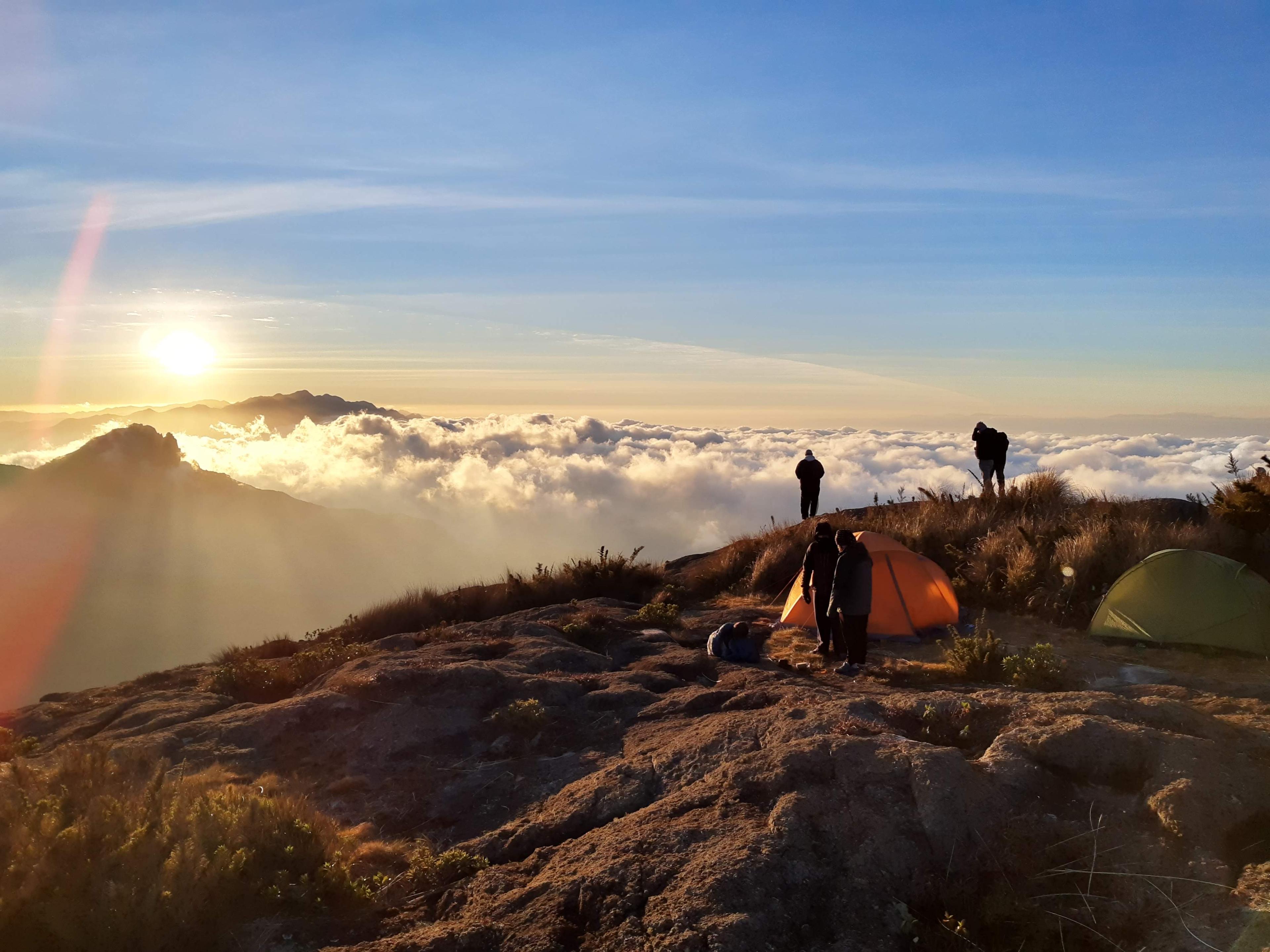 Foto tirada no nascer do sol do Pico dos Marins. Enxerga-se o sol saindo do horizonte, um mar de nuvens abaixo do pico, e algumas barracas e pessoas no primeiro plano.