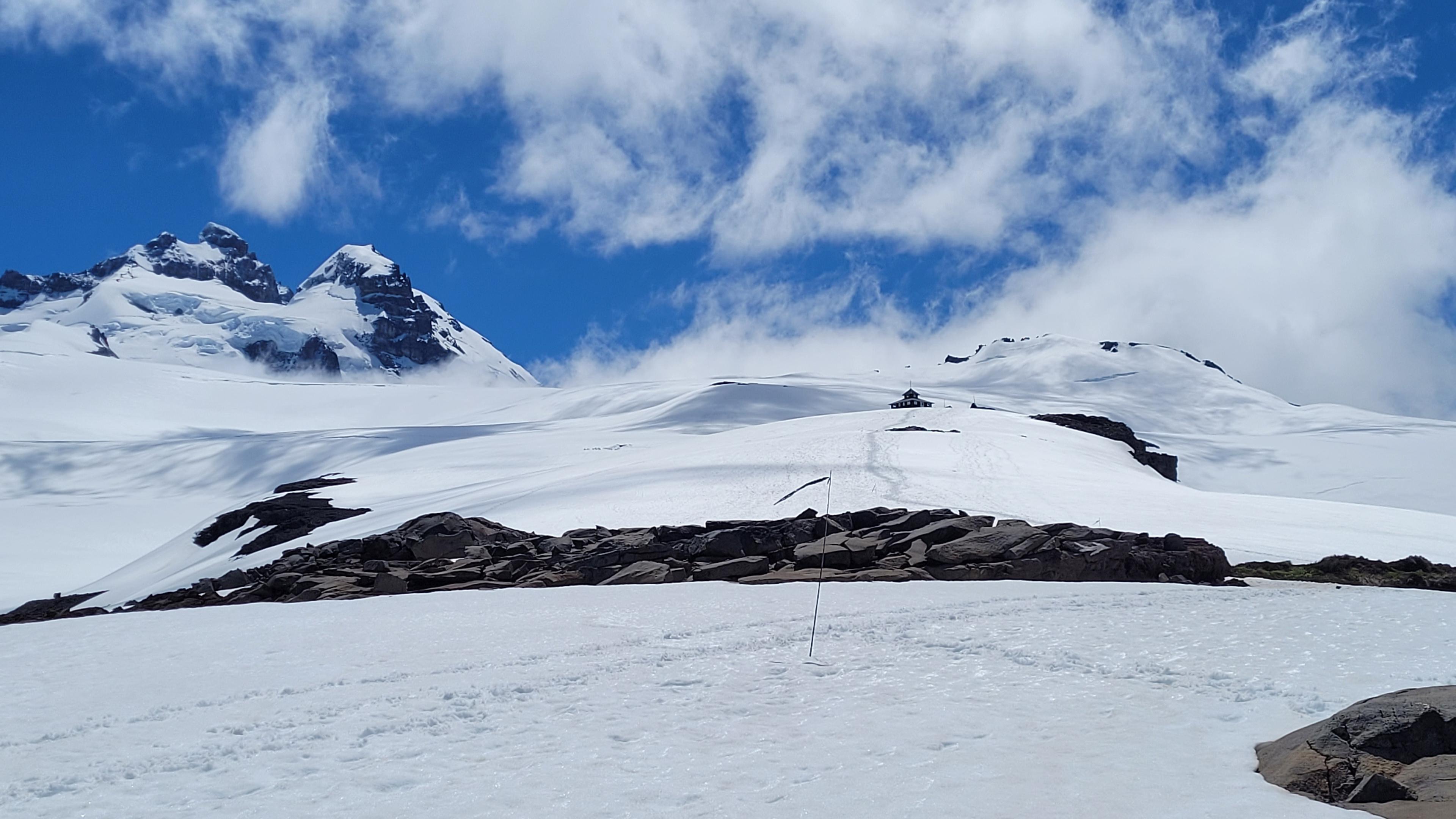 No primeiro plano, um caminho de neve e uma bandeira; no plano médio, o refúgio de montanha Otto Meiling; no último plano, o Cerro Tronador