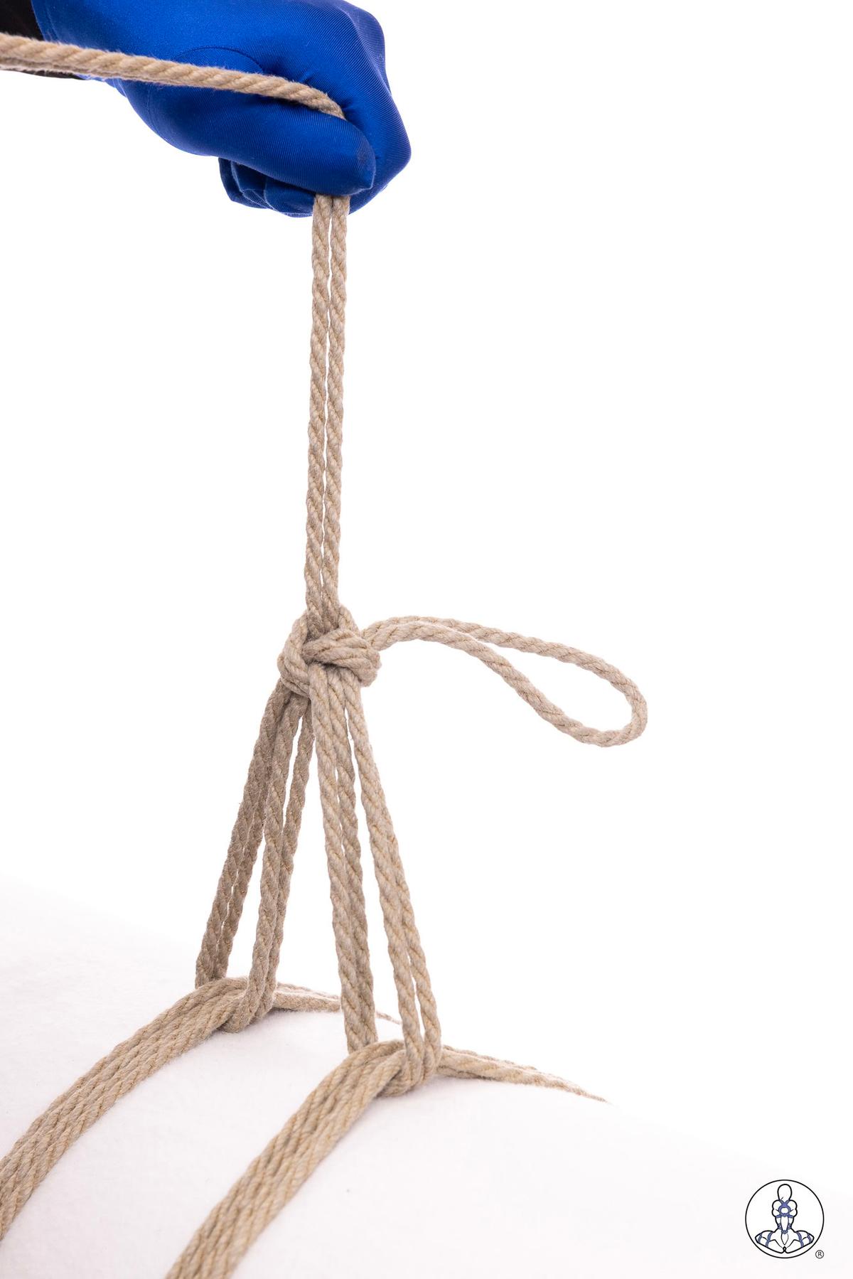 Blue-gloved hand pulling natural jute rope through a gathered bundle, demonstrating rope tension technique with multiple strands forming a loop and extending downward against white background.