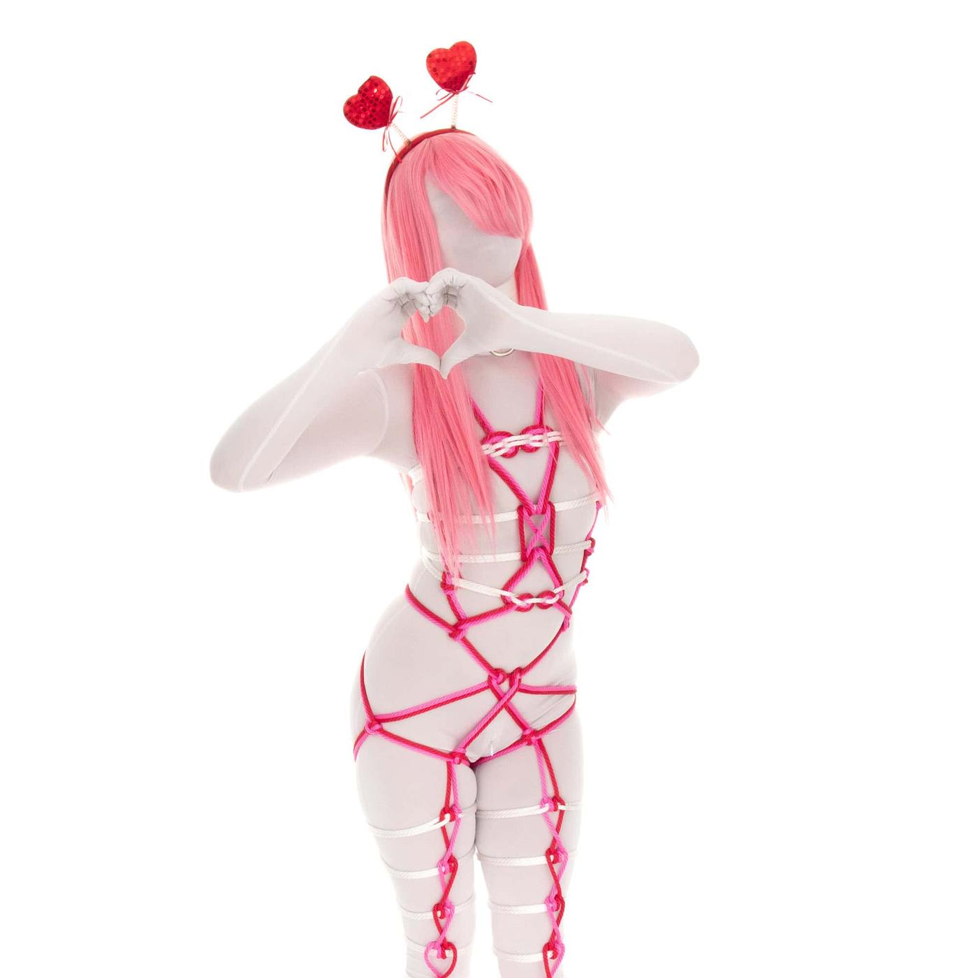 Full-body decorative rope harness in bright pink rope featuring geometric diamond patterns across torso, hips, and legs, with white rope accents on upper chest and thighs. Figure stands with arms raised forming heart shape with hands.
