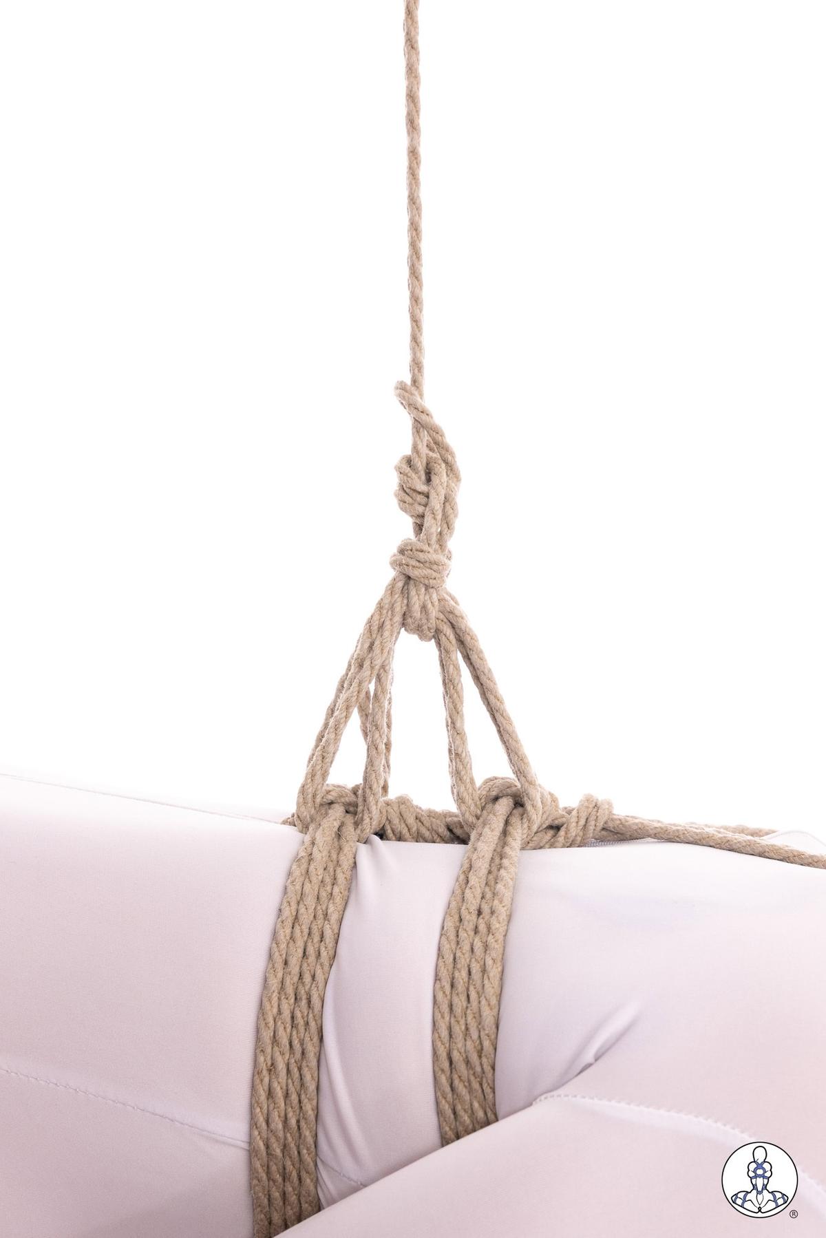 Natural jute rope suspension harness around white cushion showing basic hip harness configuration with vertical support lines and gathered knot at top. The rope forms two parallel bands across the cushion connected to an overhead suspension point.