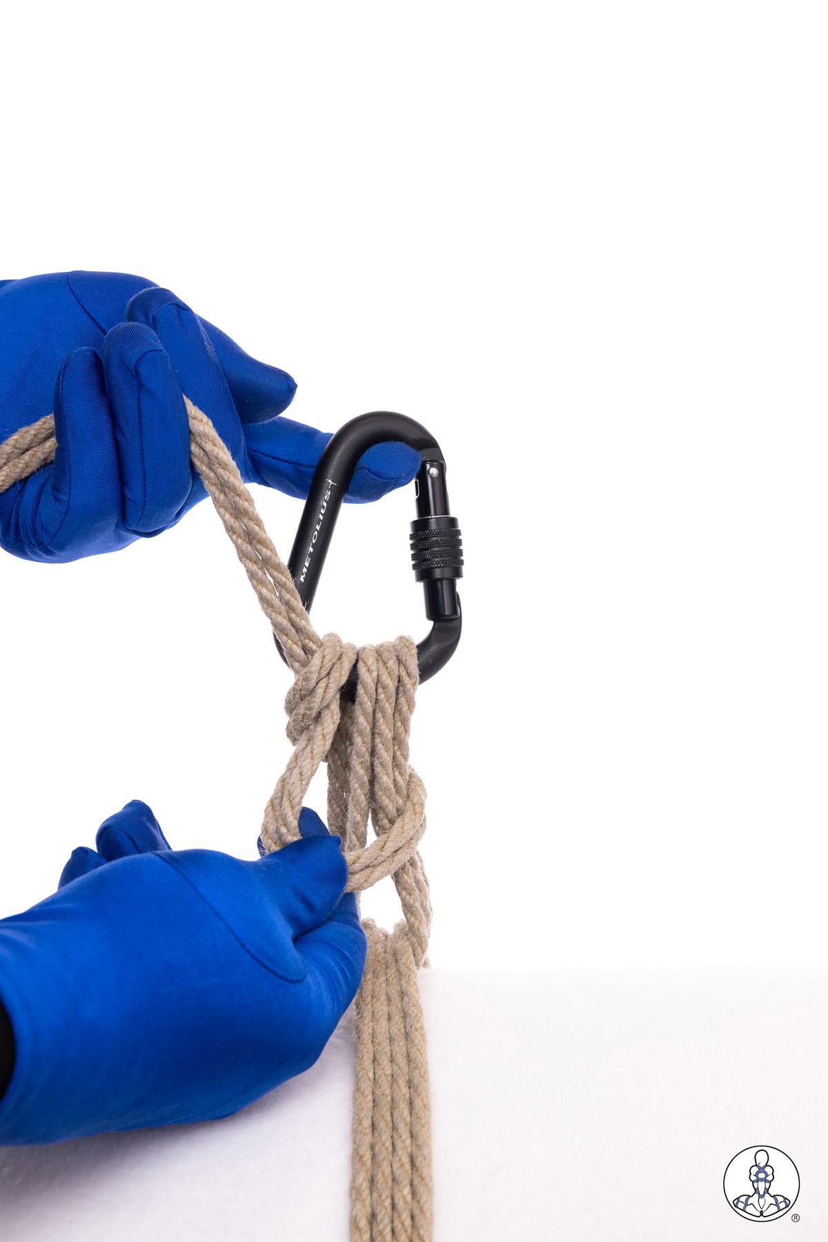 Blue-gloved hands threading natural jute rope through a black locking carabiner, demonstrating rope attachment or rigging technique against white background.