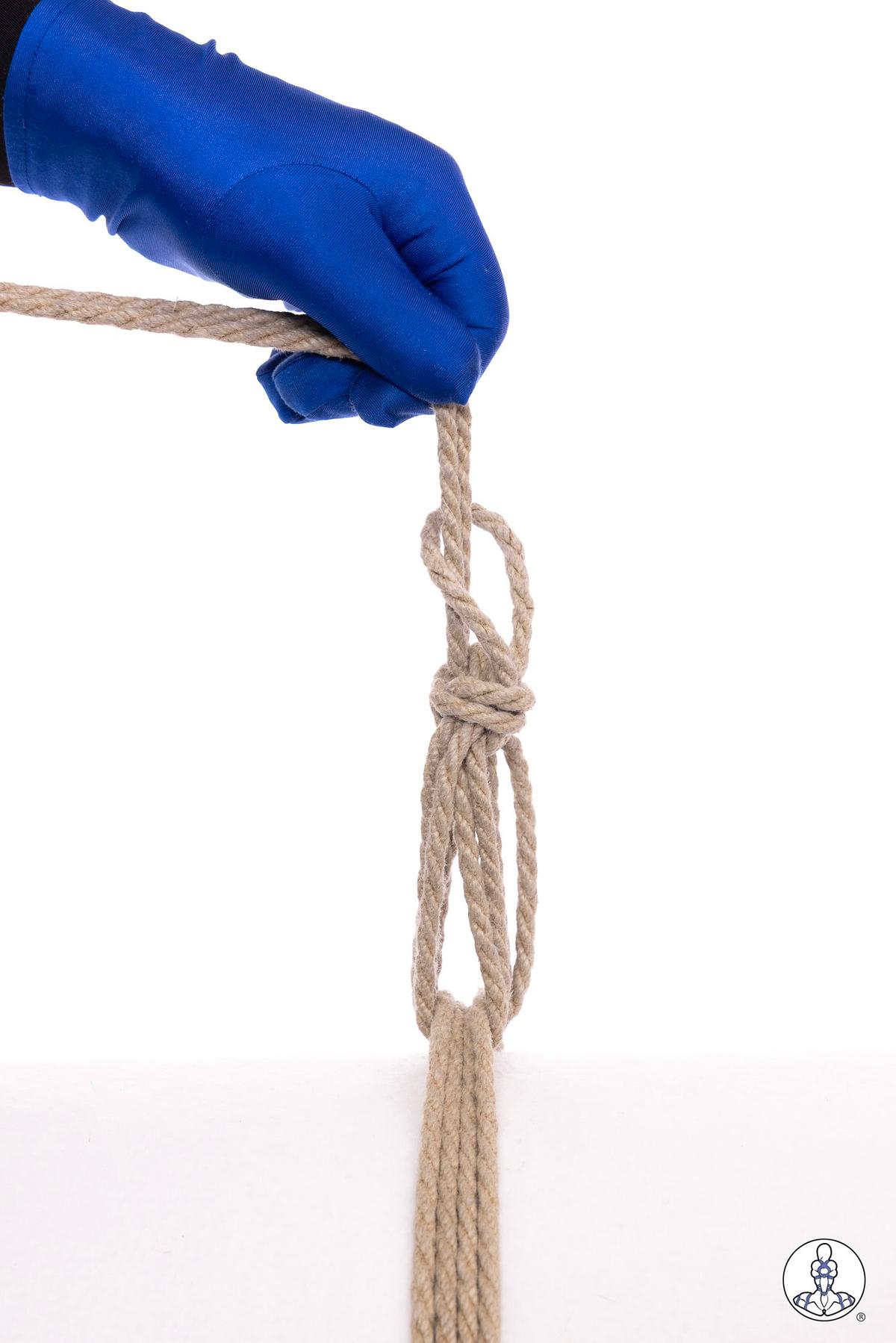 Blue-gloved hand pulling natural jute rope through a partially completed overhand knot with multiple rope strands, demonstrating a bundling or gathering technique against white background.