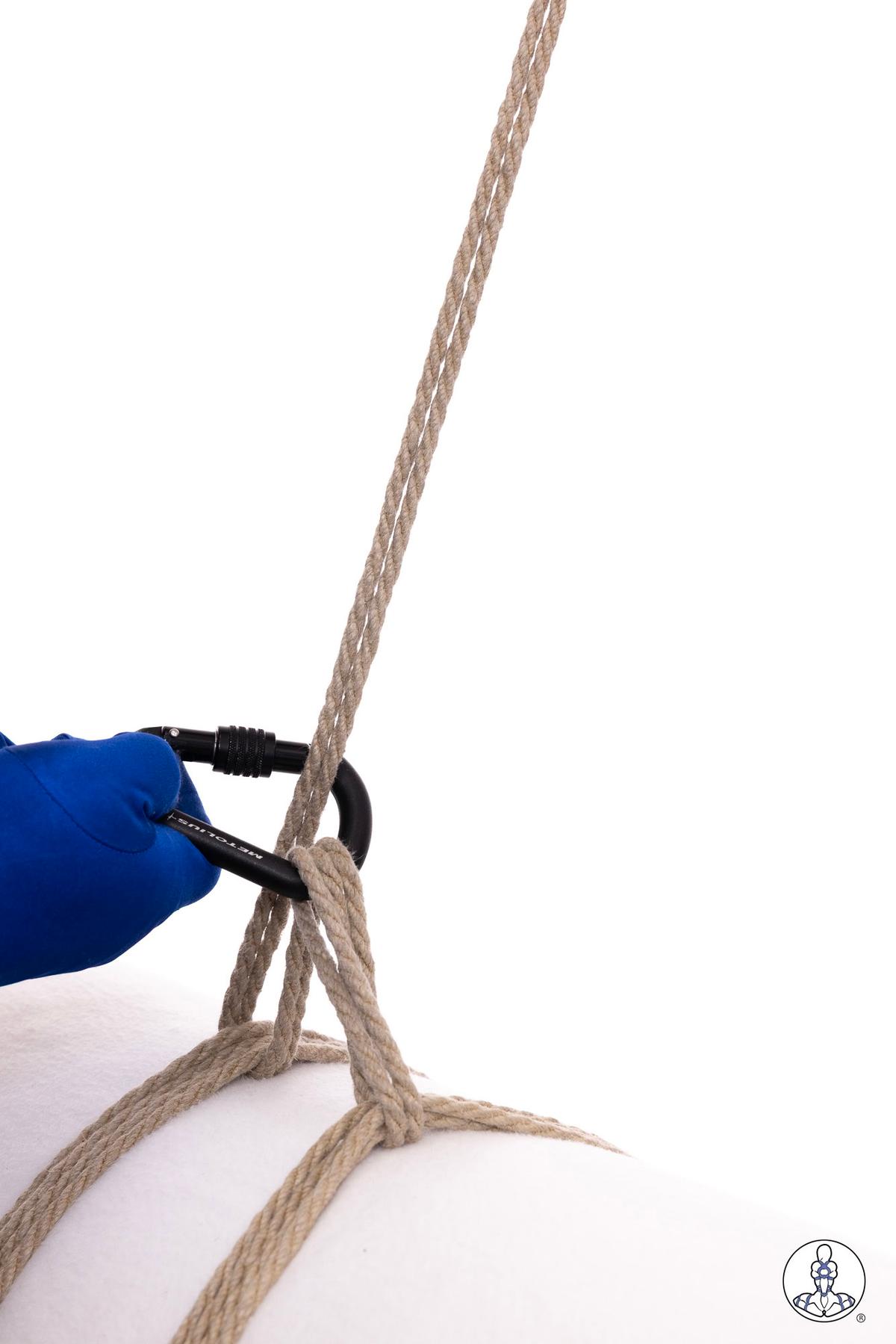 Blue-gloved hand using black carabiner to thread natural jute rope through a partially completed knot configuration, demonstrating rope manipulation technique on white surface.