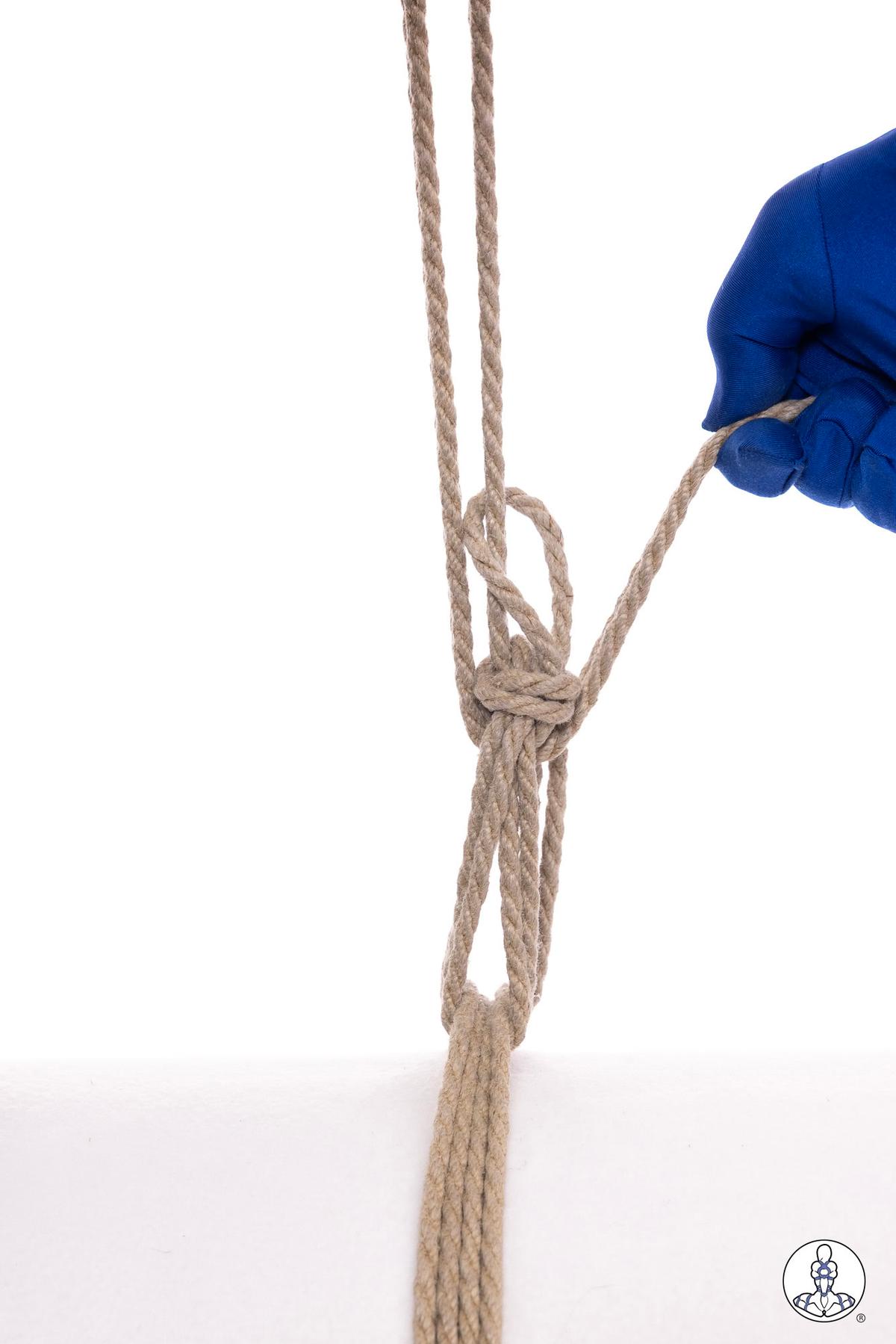 Blue-gloved hand holding natural jute rope demonstrating a munter hitch knot formation with doubled rope strands against white background.