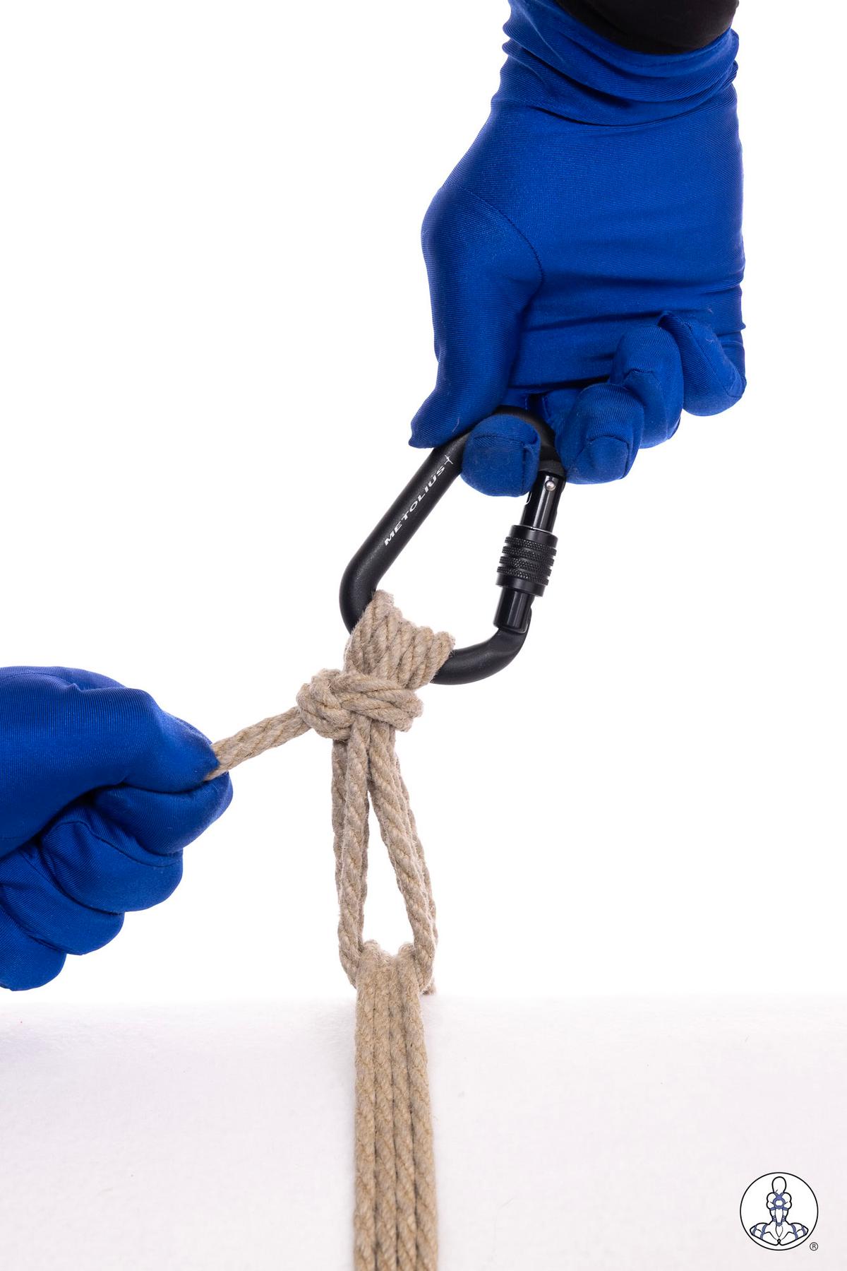 Two blue-gloved hands threading natural jute rope through a black metal carabiner hook, demonstrating rope preparation or rigging technique against a white background.