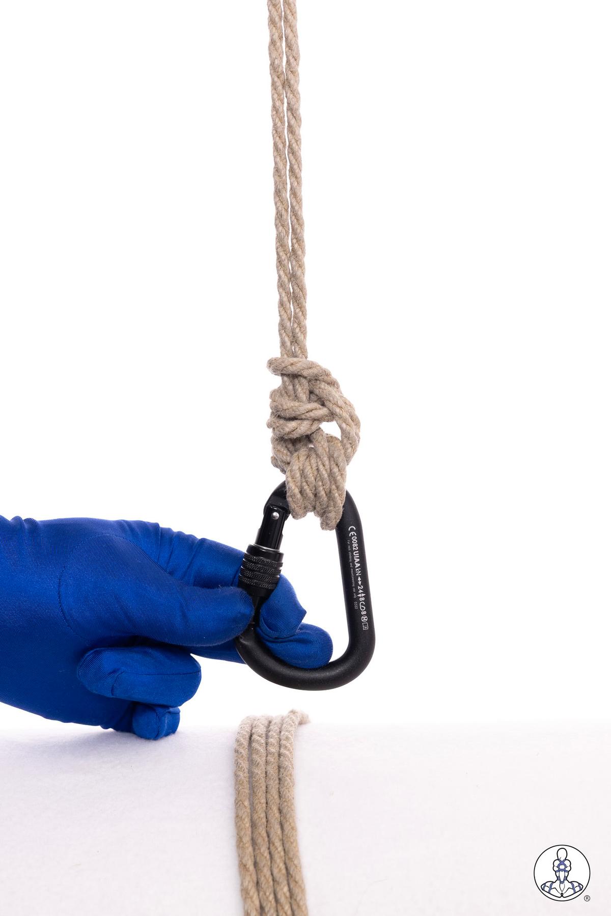 Natural jute rope threaded through a black carabiner, held by a blue-gloved hand, demonstrating a suspension rigging attachment point with visible rope wraps around the carabiner.