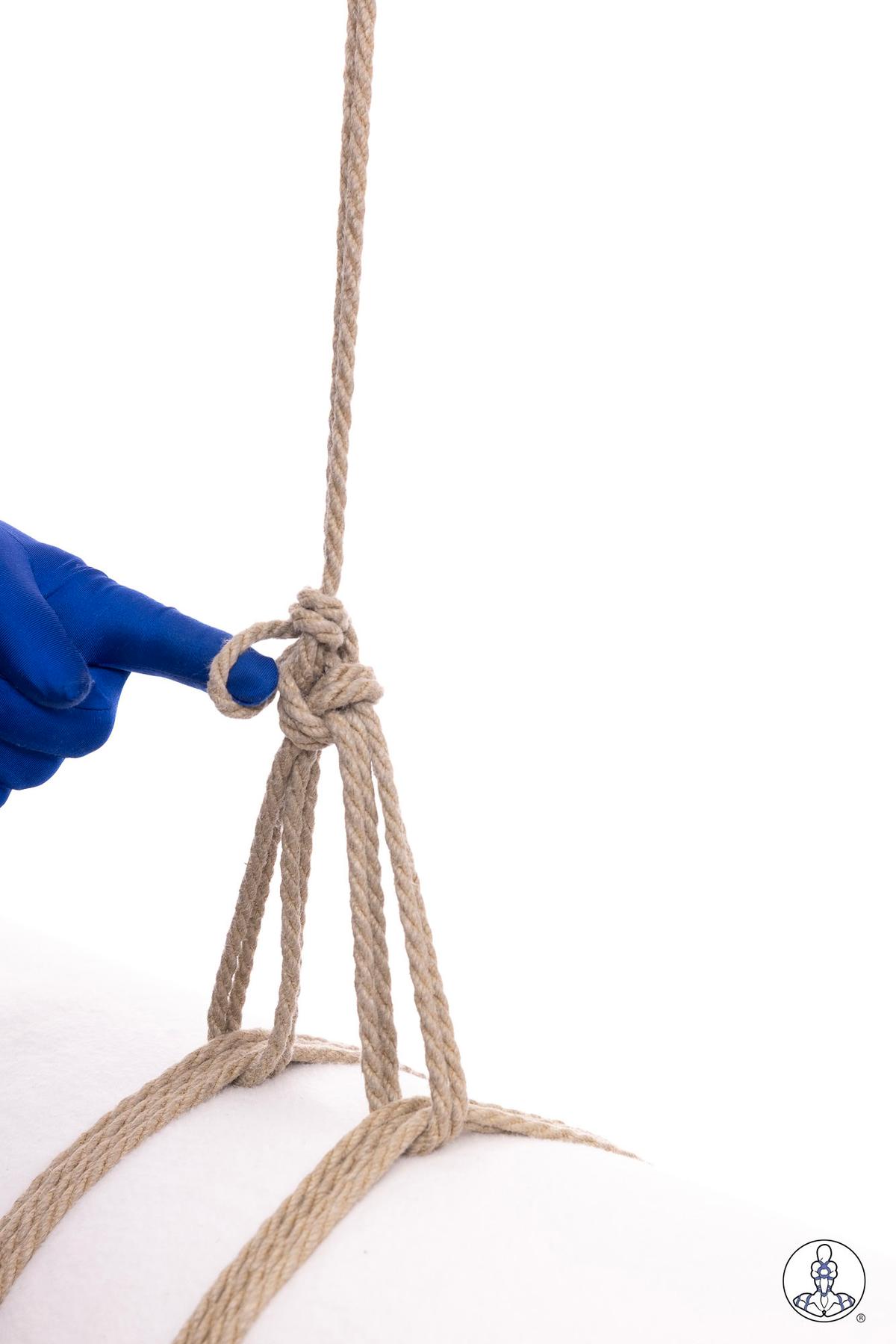 Blue-gloved hand pointing to a gathering knot in natural jute rope, showing four strands bundled together below a vertical suspension line with loose rope ends extending downward.
