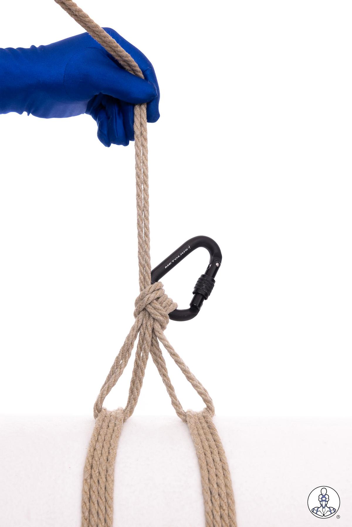 Blue-gloved hand holding natural jute rope threaded through a black carabiner, demonstrating rope handling technique with two rope strands forming loops below the carabiner.