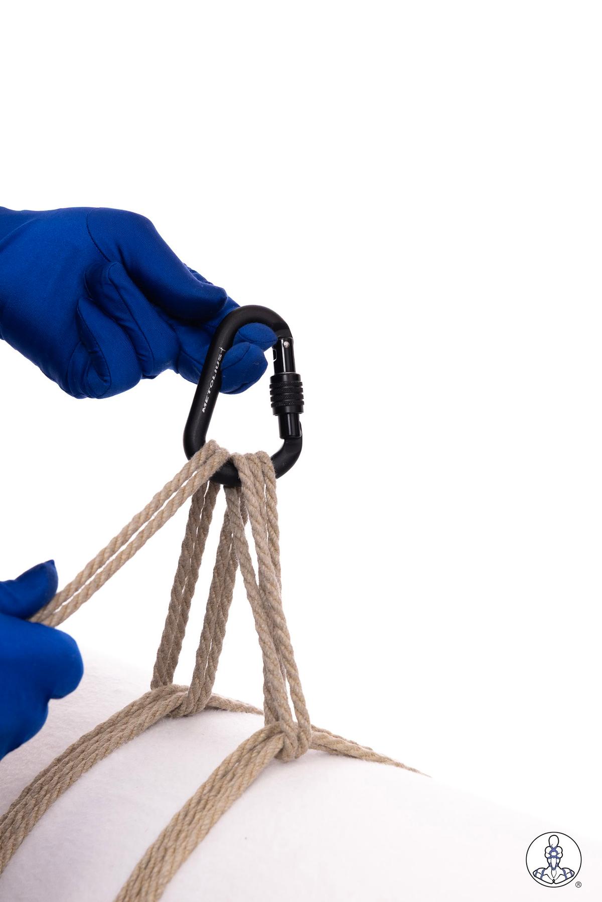 Blue-gloved hands threading natural jute rope strands through a black carabiner clip, demonstrating rope preparation or rigging technique against a white background.