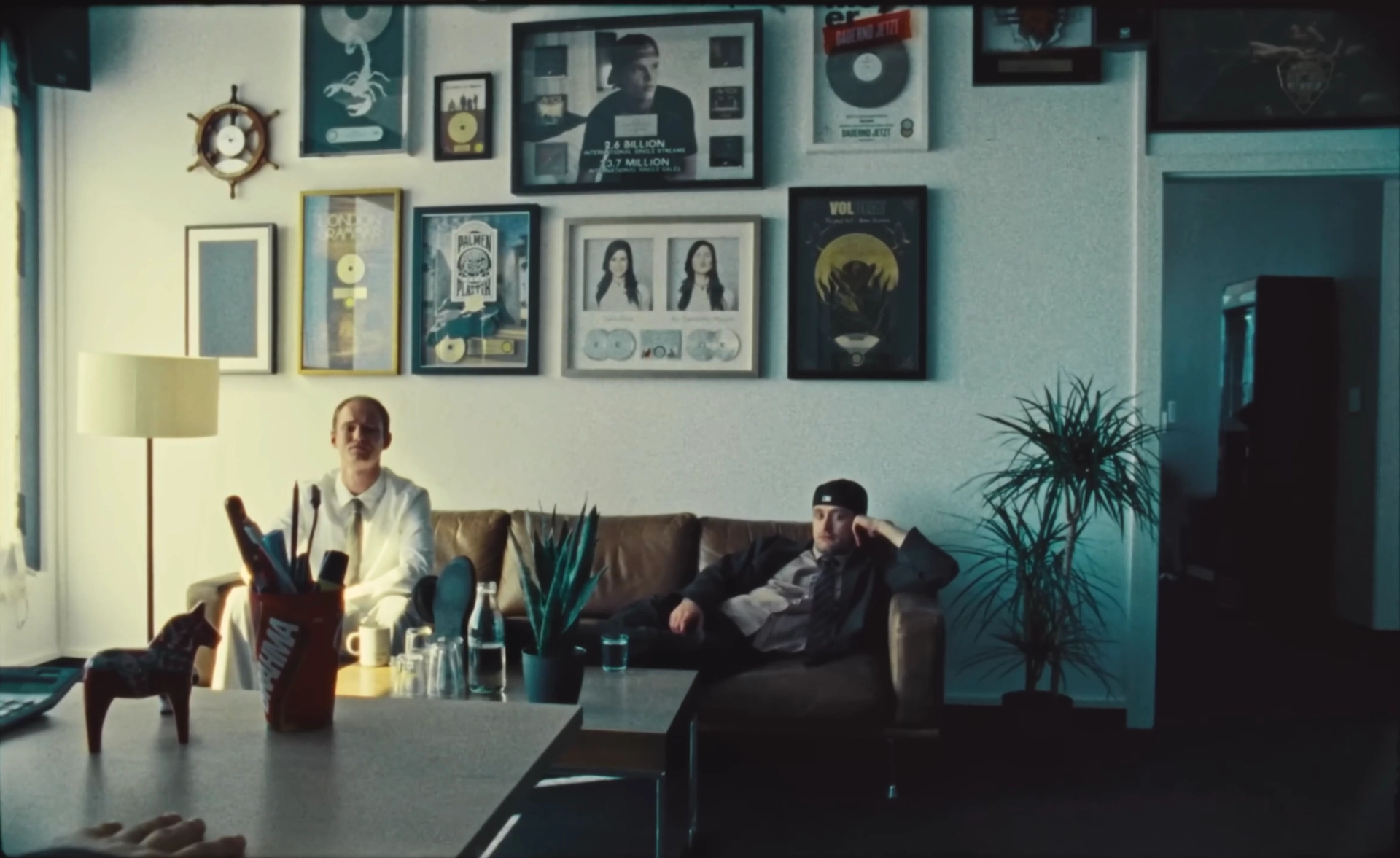 The waiting-room shot without the inset: both men slouch on the sofa, seen across a desk with a painted wooden horse and a cup of pens.