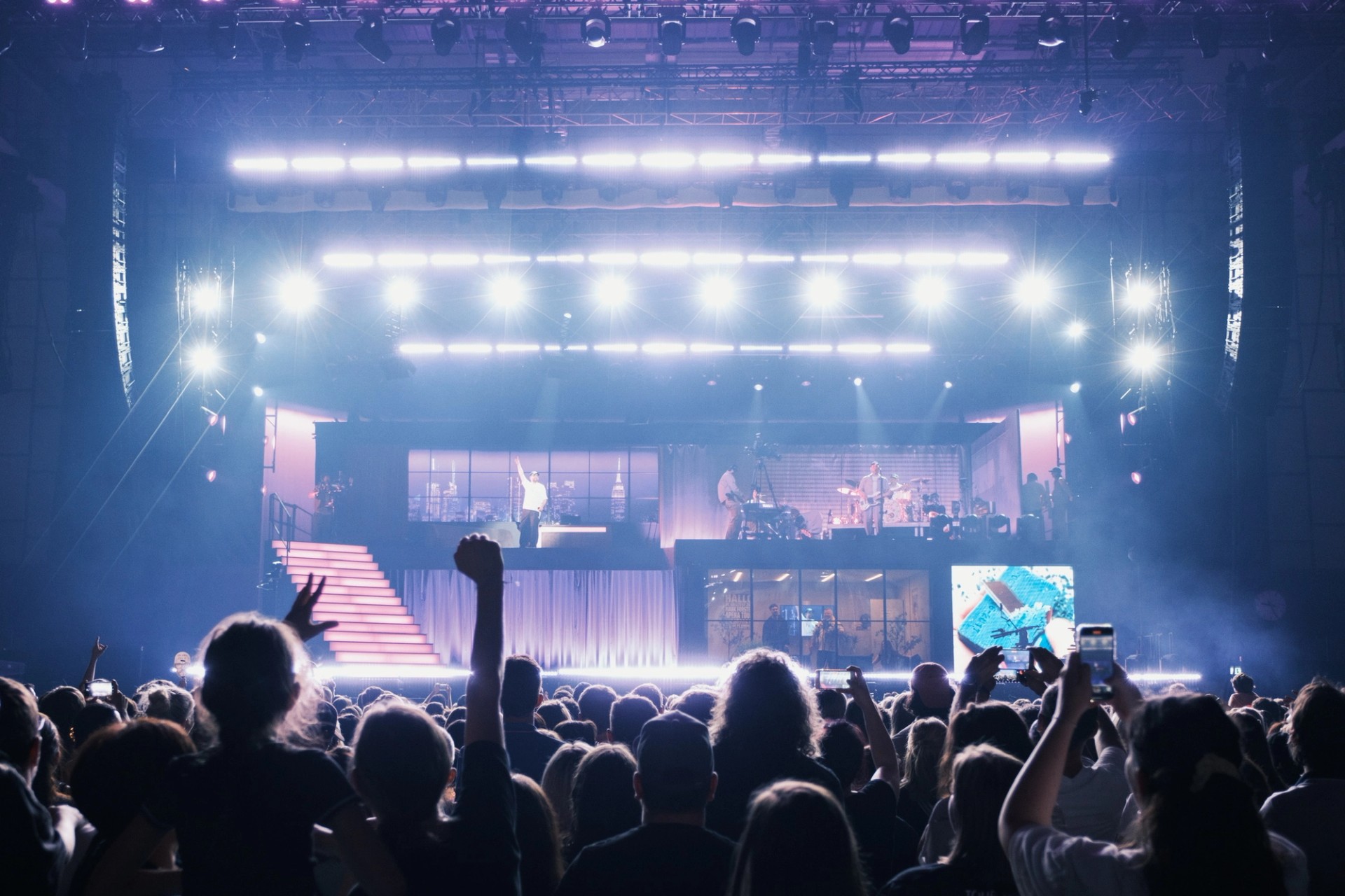 The stage in blue-white light seen from within the crowd, fans raising arms and phones.