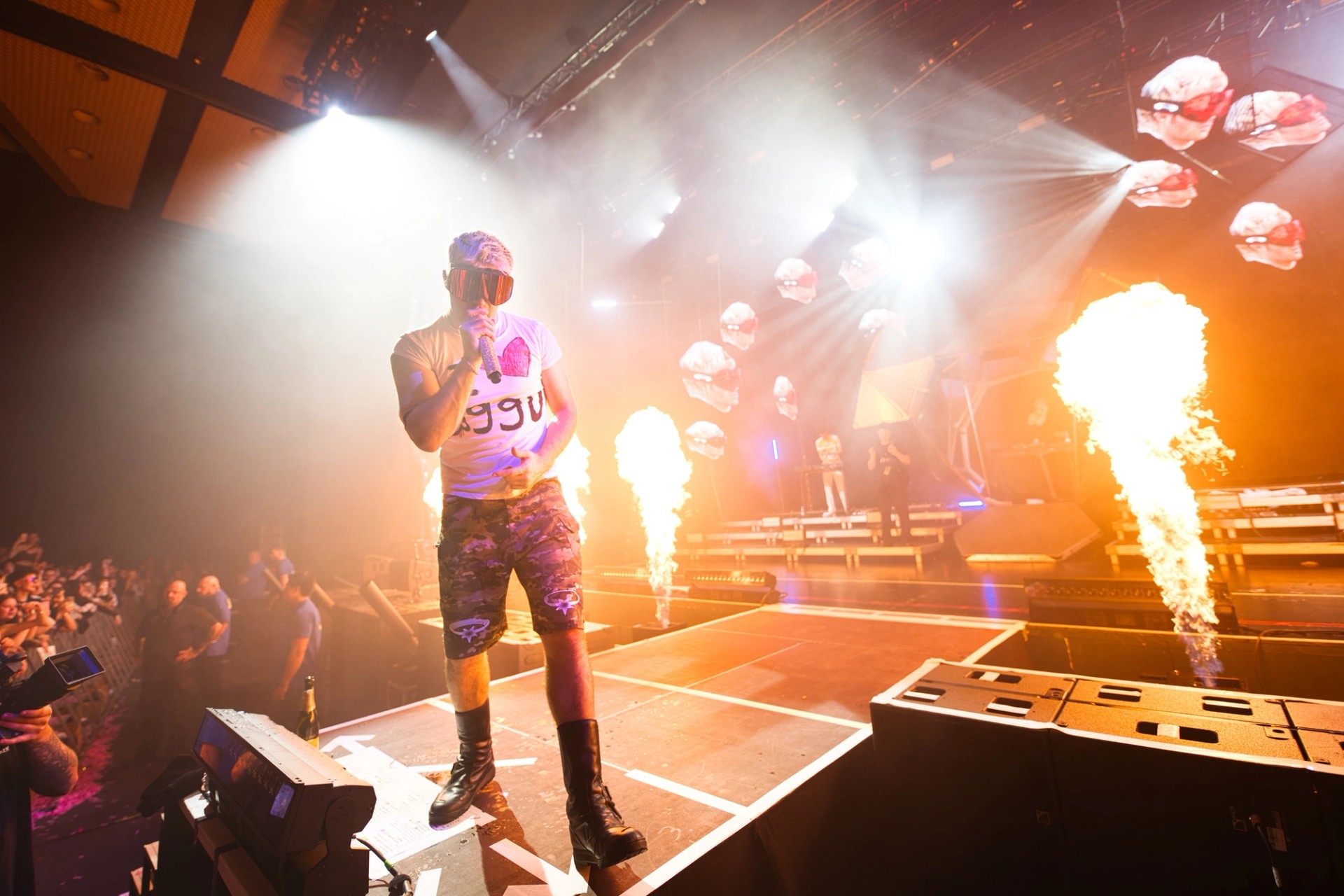 Ski Aggu at the front of the stage in a white T-shirt and camo shorts, flame jets firing beside him and LED panels showing heads in red goggles.