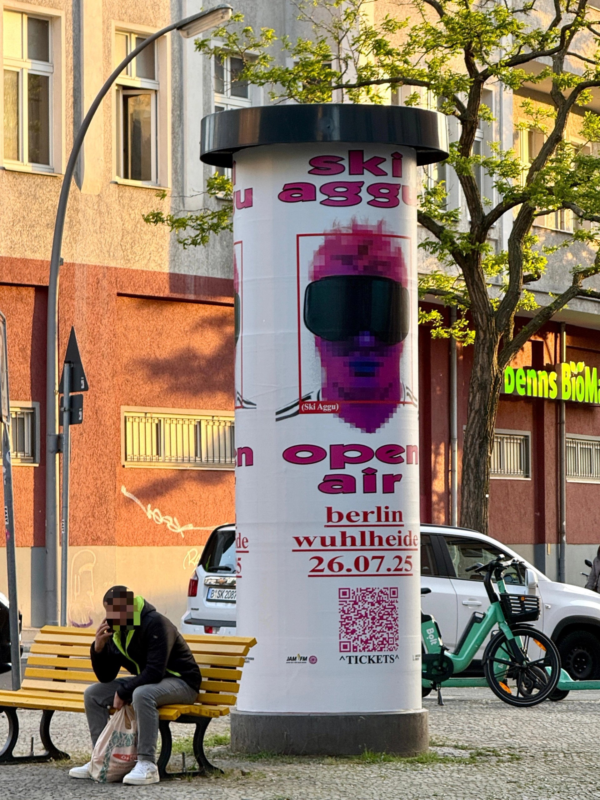 Advertising column on a Berlin street wrapped in the Ski Aggu open air poster for Wuhlheide on 26.07.25, a person sitting on a yellow bench beside it.